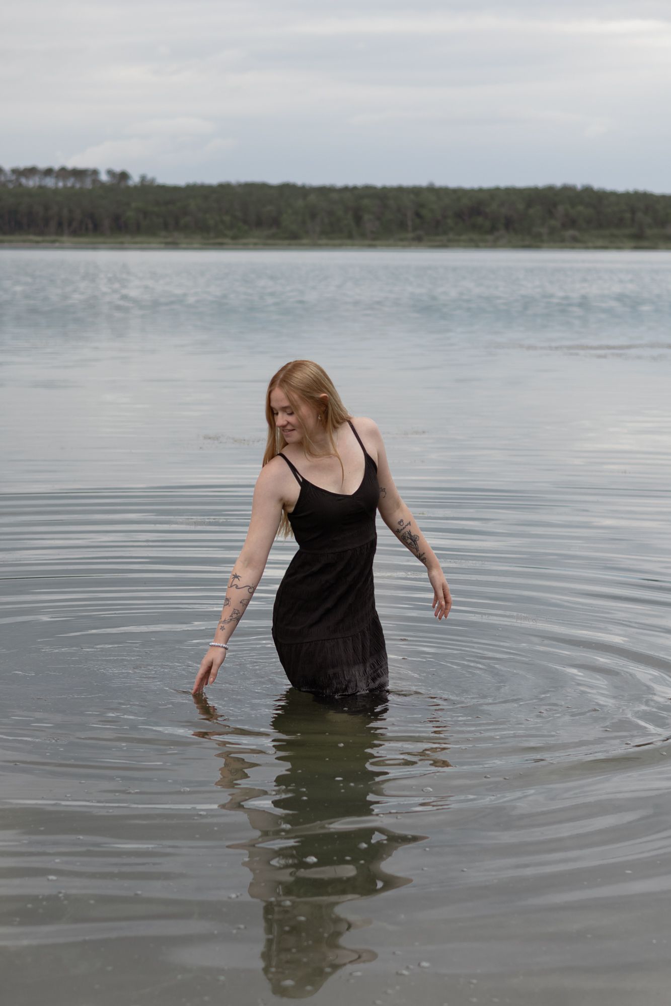 A tattooed redhead woman in a black dress wading in a calm lake with a forested shoreline in the background.
