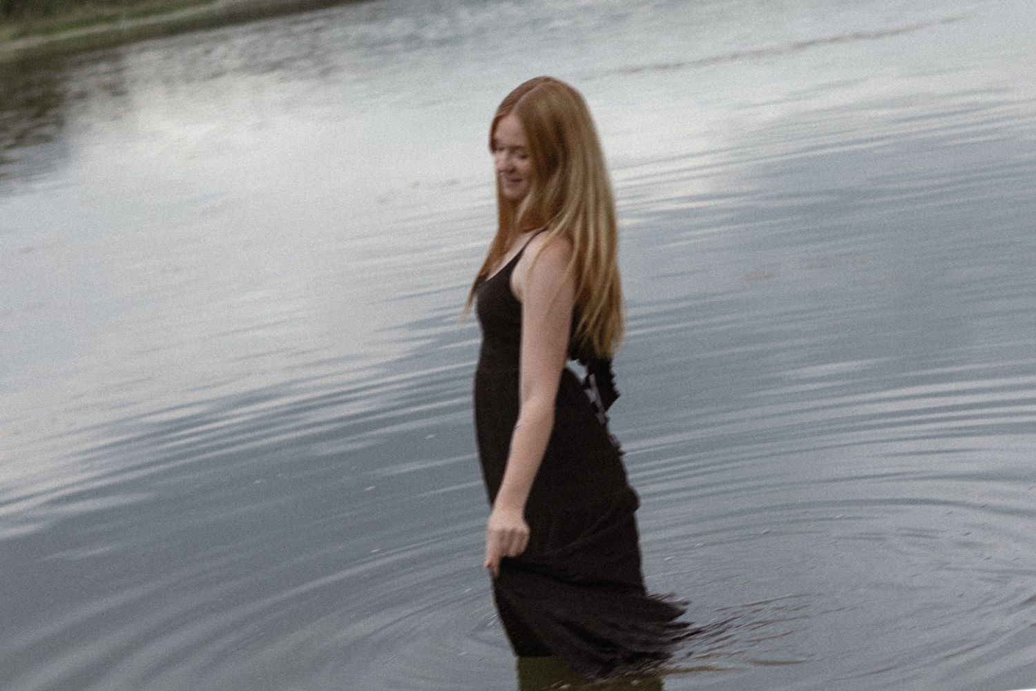 Young woman with long red hair standing in a lake wearing a black dress, water up to her knees.