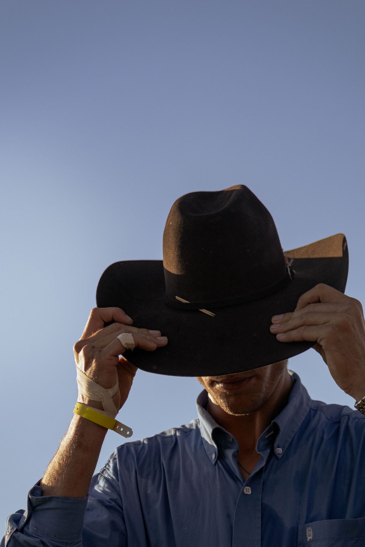 Man in blue shirt adjusting dark brown cowboy hat against clear blue sky, wearing yellow wristband.