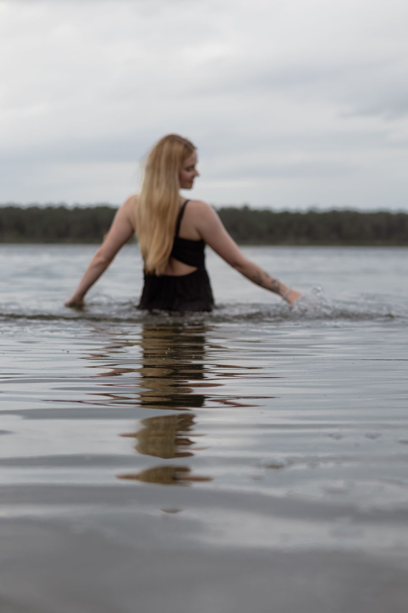A blonde woman in a black swimsuit wading through calm lake water on an overcast day.