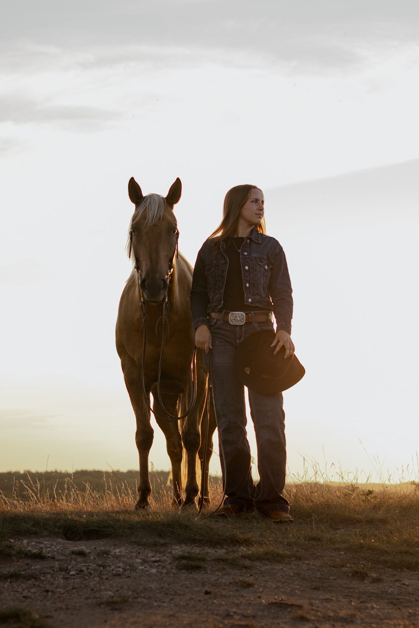 Young cowgirl with long hair holds a hat beside a chestnut horse at golden sunset in an open field.