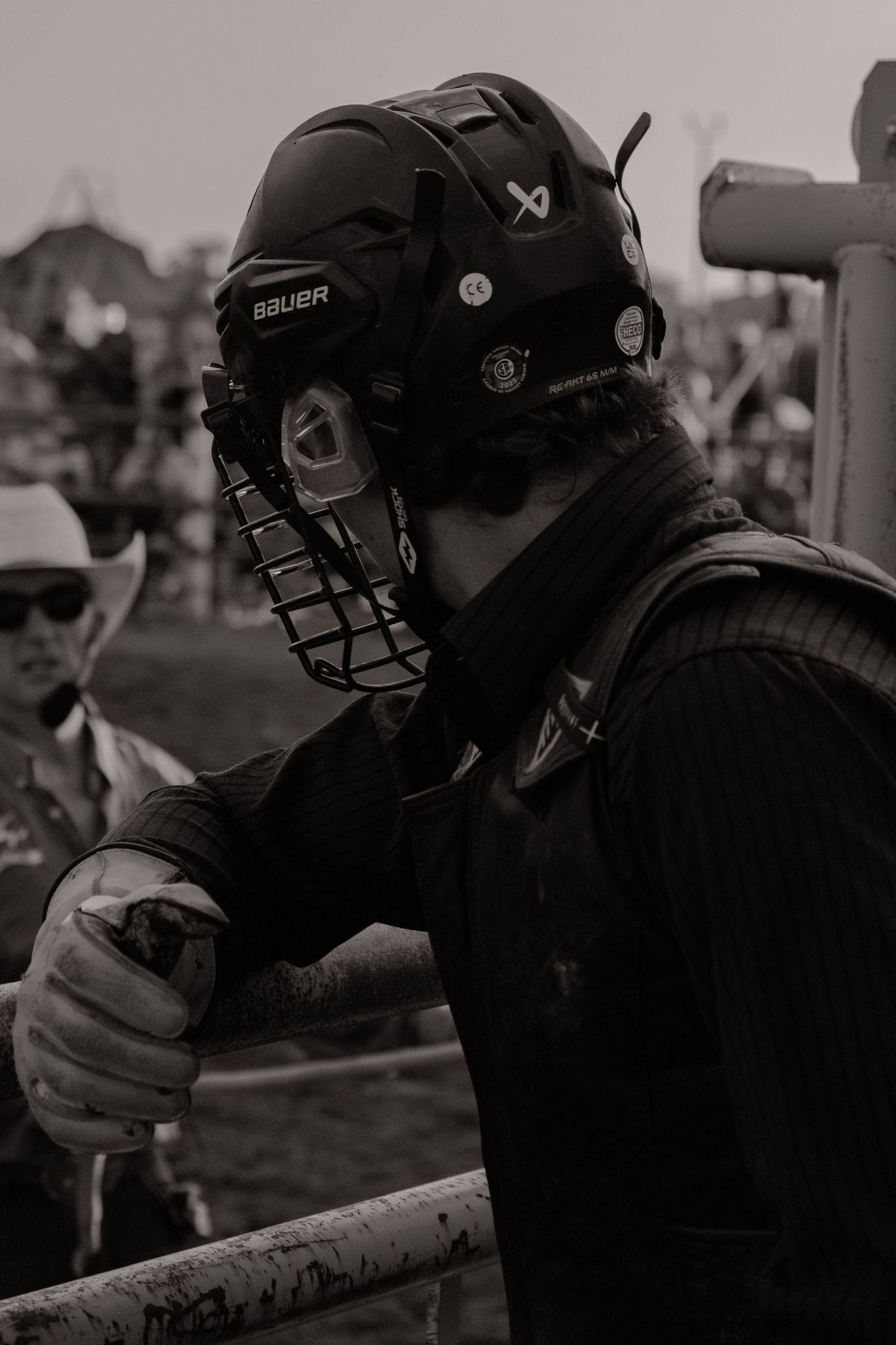 Black and white photo of a person wearing a Bauer hockey helmet with cage at an outdoor rodeo event.