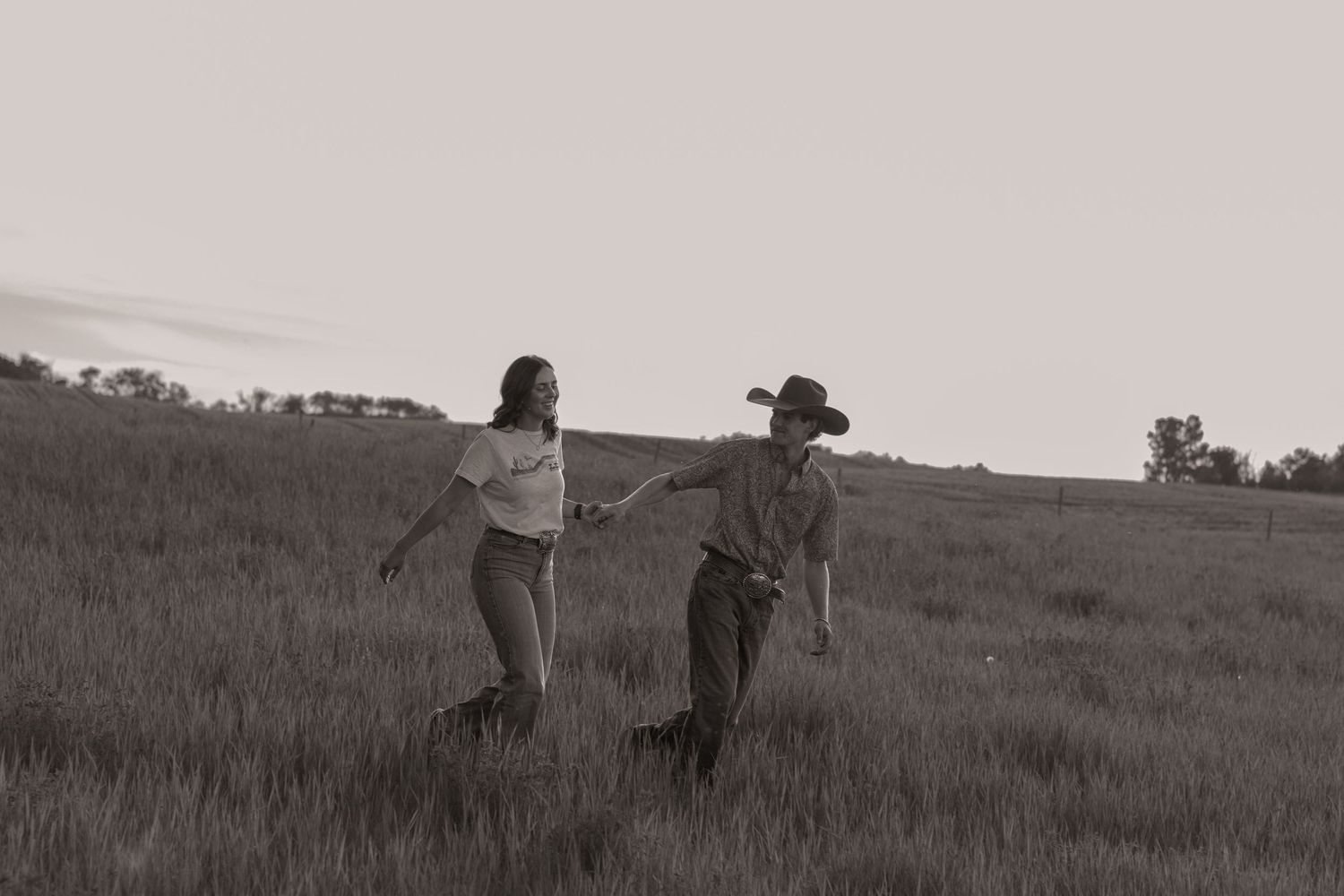 A couple holds hands walking through a grassy field at dusk in a black and white countryside photo.