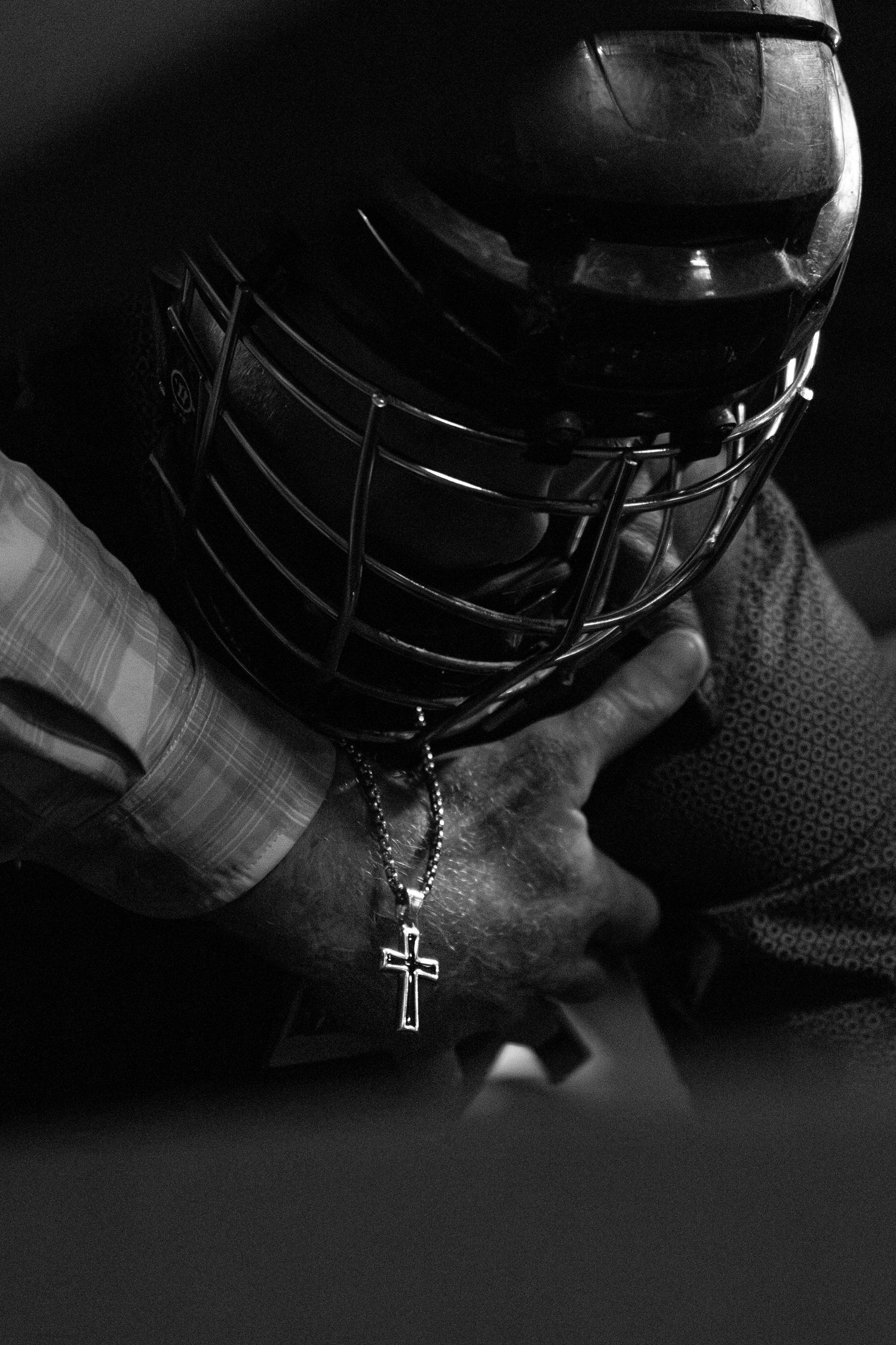 Black and white photo of a football player in helmet holding a cross necklace, bowing head in prayer.