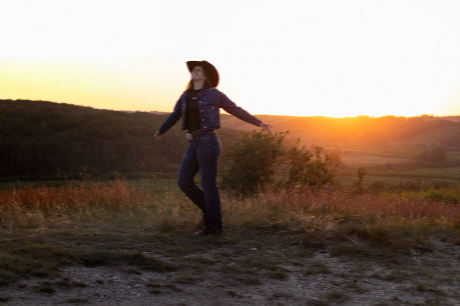 A person in a cowboy hat dances at golden sunset in a scenic rural landscape with rolling hills.