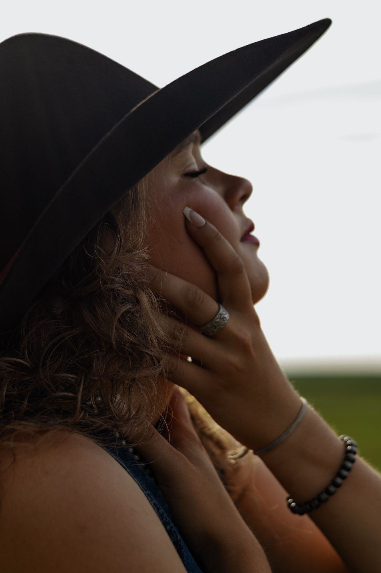 Woman in black wide-brim hat with eyes closed, hands gently touching her face, wearing rings and a beaded bracelet.