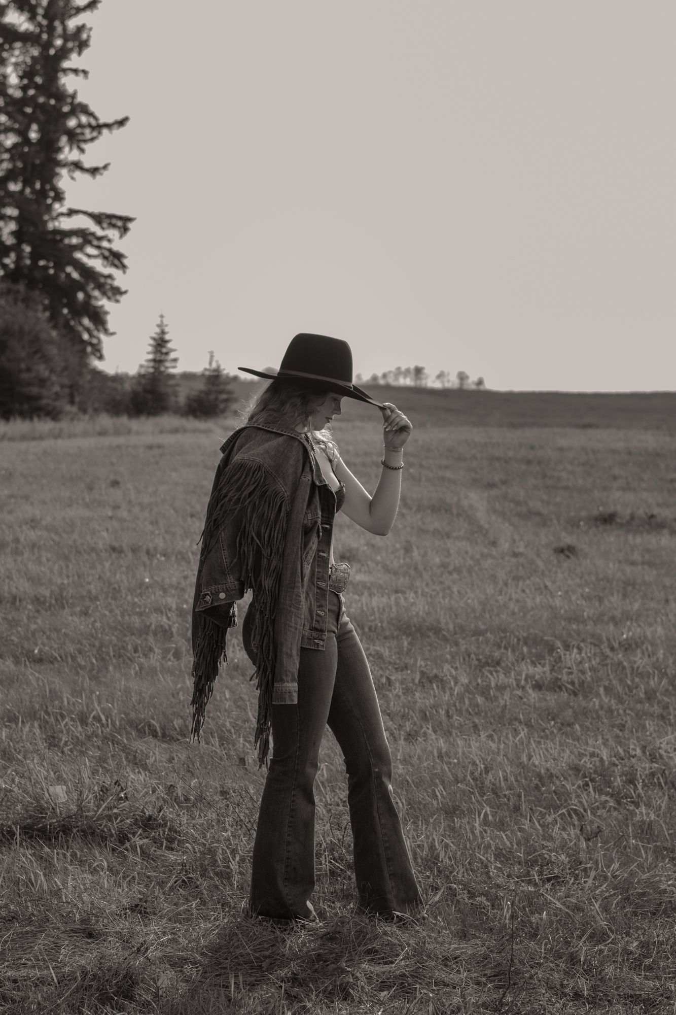 Woman in fringed vest and black cowboy hat standing in field, black and white photo.