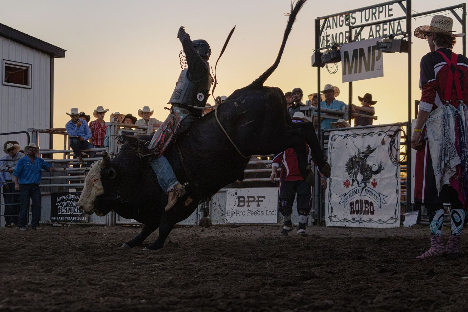 A rodeo cowboy rides a bucking bull at sunset during a professional bull riding competition at an outdoor arena.