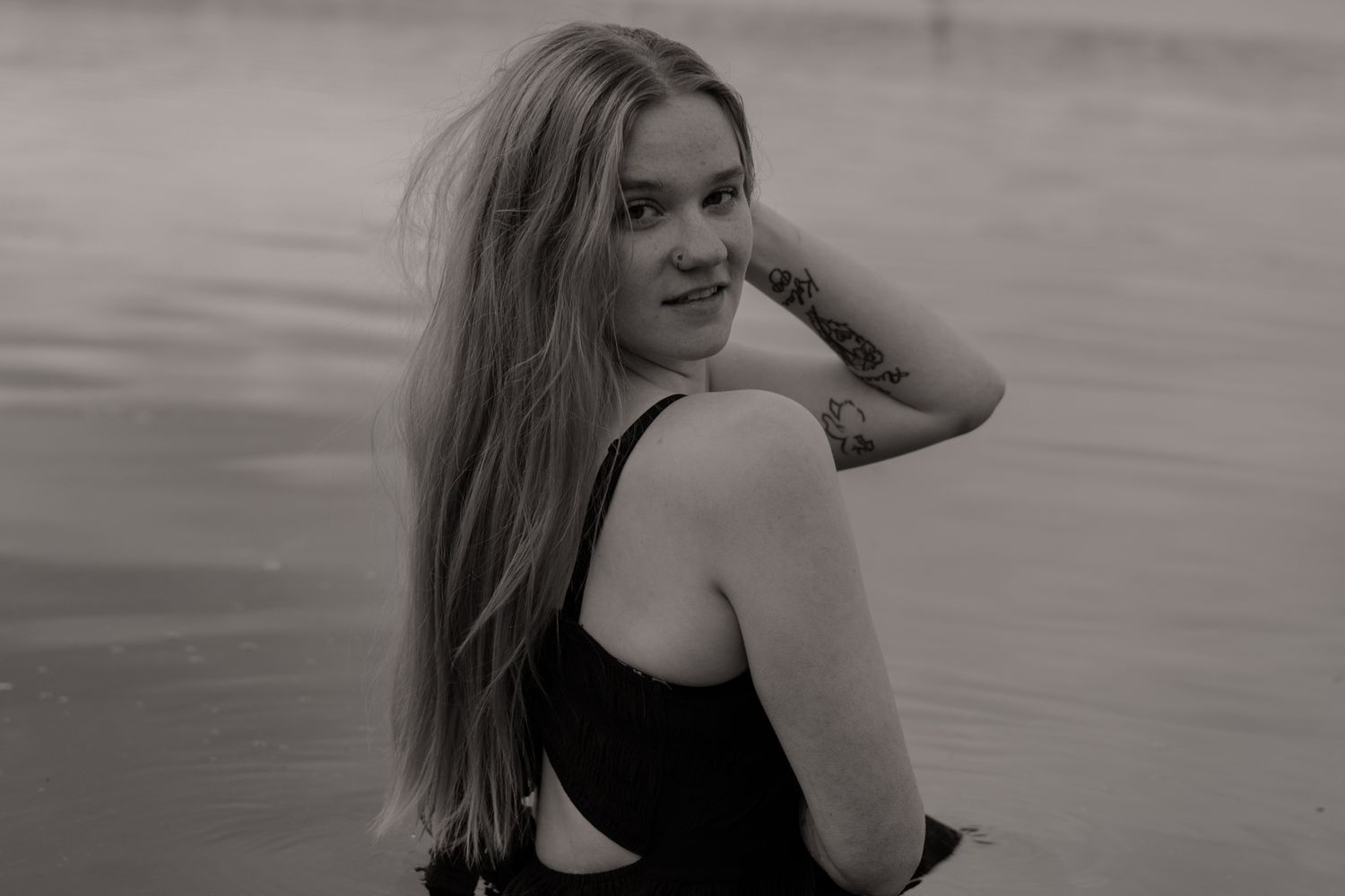 Black and white photo of a young woman with long hair and tattoo sitting by the water, smiling over her shoulder.