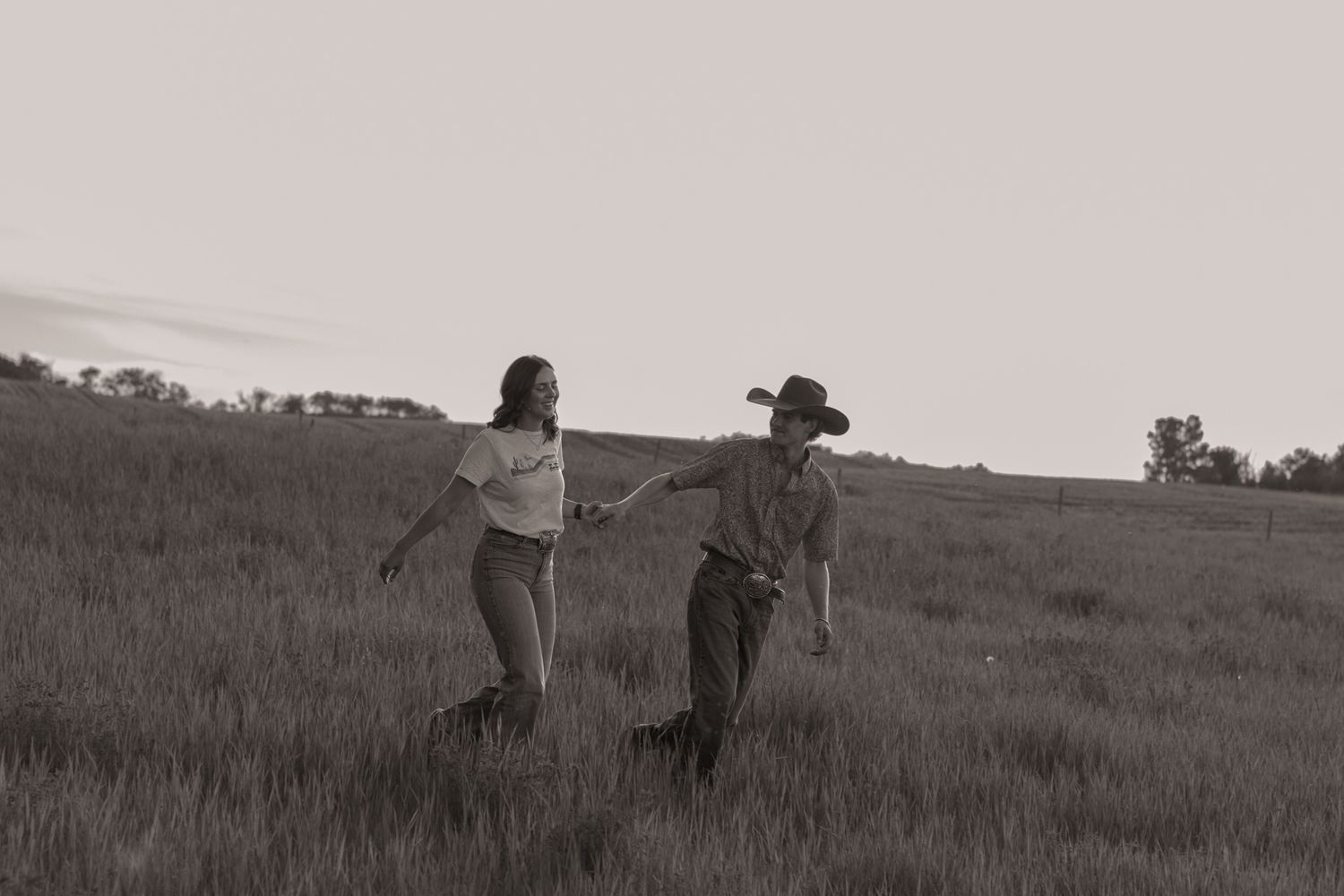 A couple walks hand-in-hand through a grassy field at dusk, the man wearing a cowboy hat, in a black and white photo.