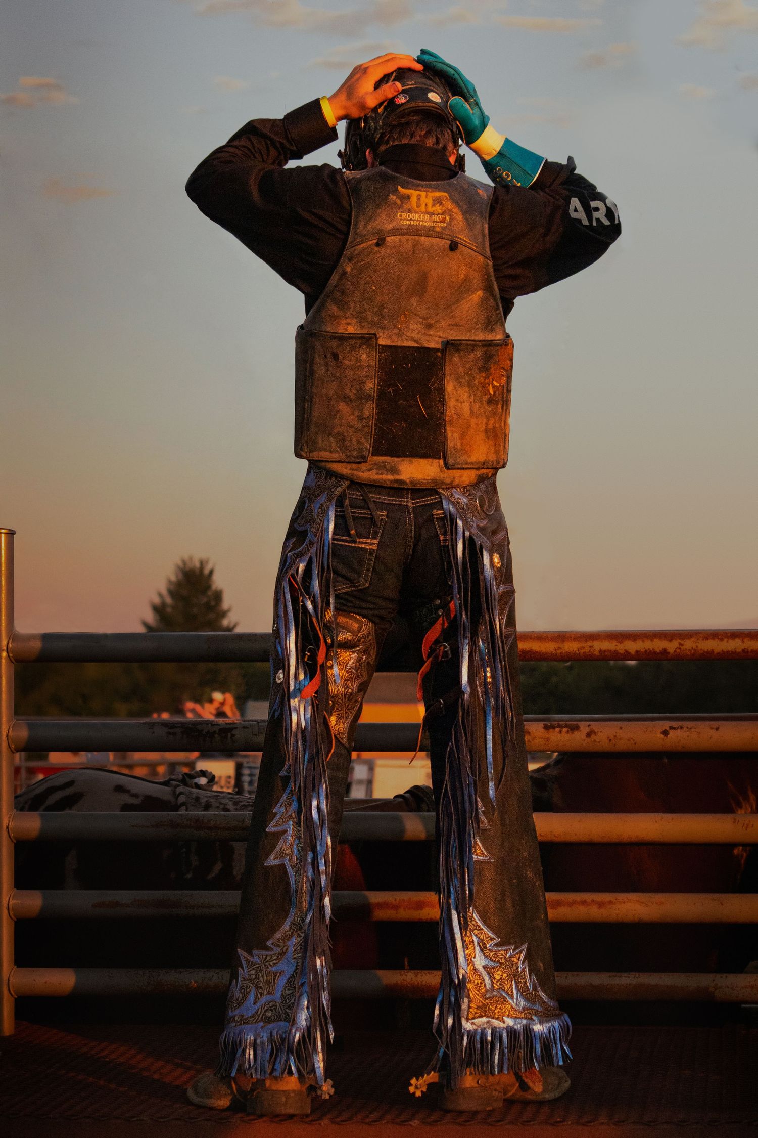 A bull rider in protective vest and shiny silver chaps adjusts helmet at sunset golden hour rodeo arena.
