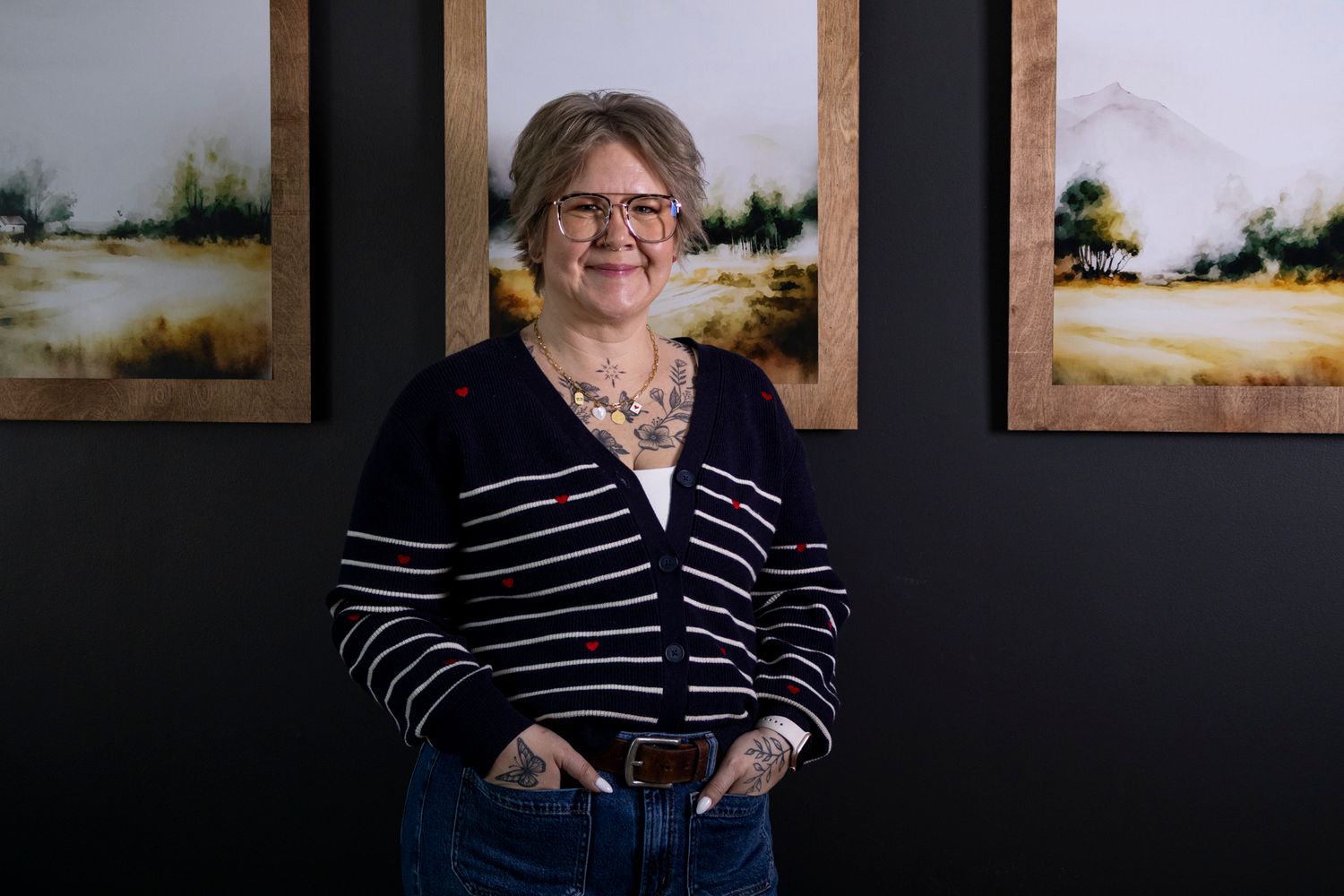 Woman in striped cardigan smiling, standing before landscape paintings on dark wall, showcasing tattoos and casual style.