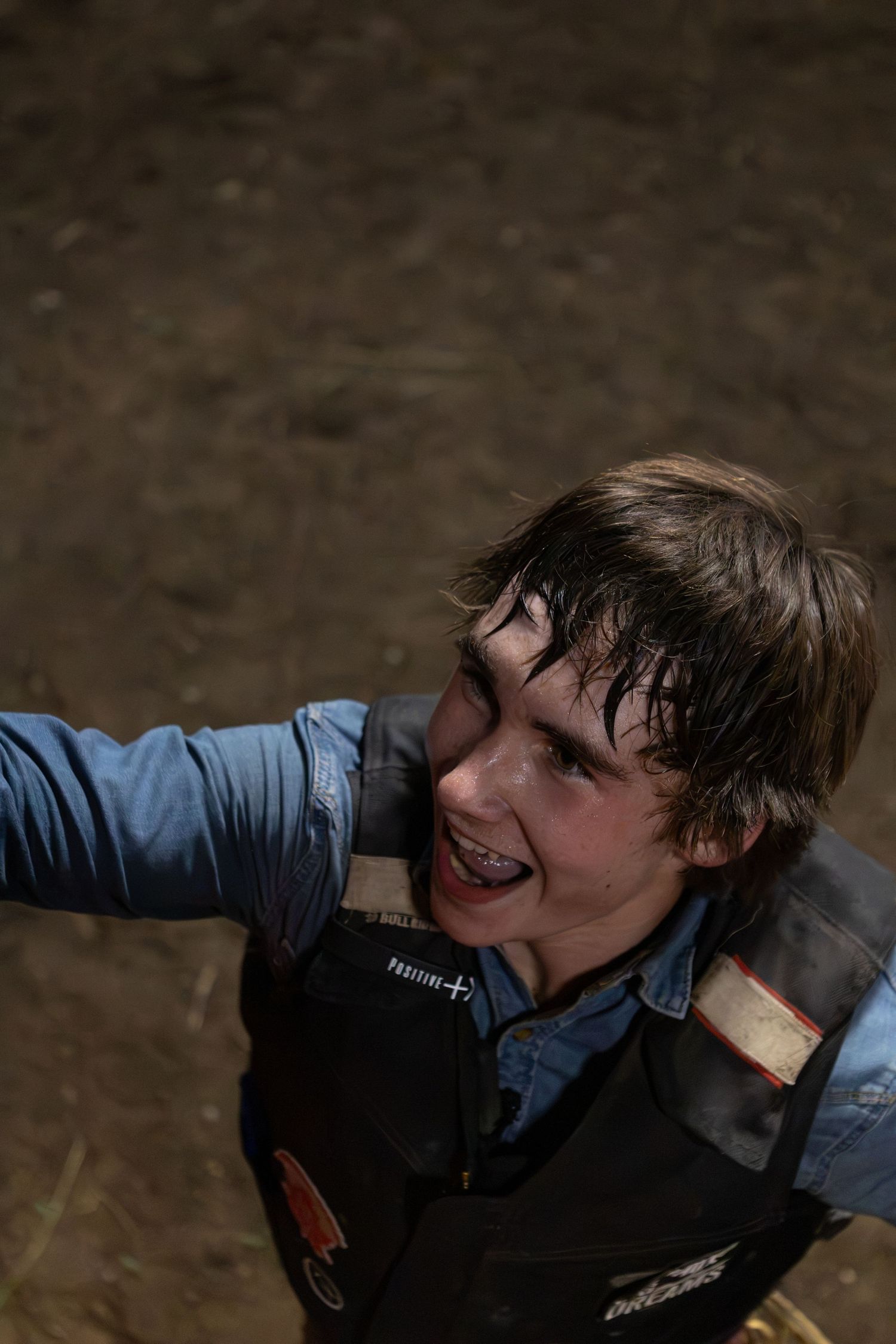 Young boy with braces and wet hair, wearing a blue shirt and black vest, looking up outdoors.
