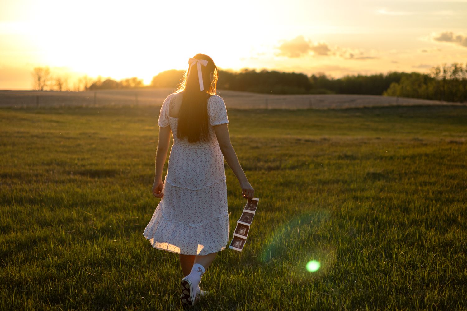 Young woman in a white dress holding a guitar walking through a green field at golden sunset.