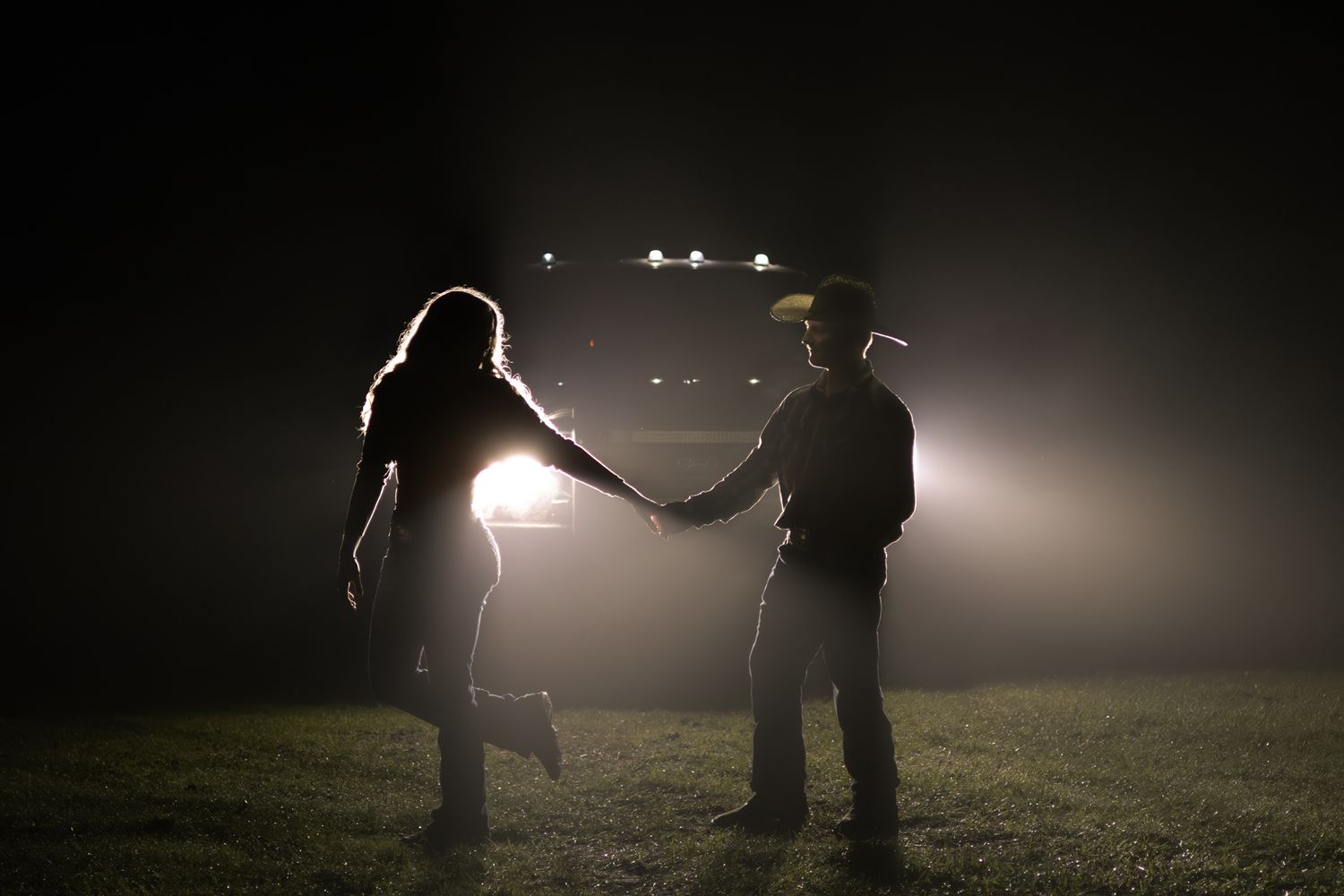 Couple holding hands silhouetted against bright headlights on a dark field at night.