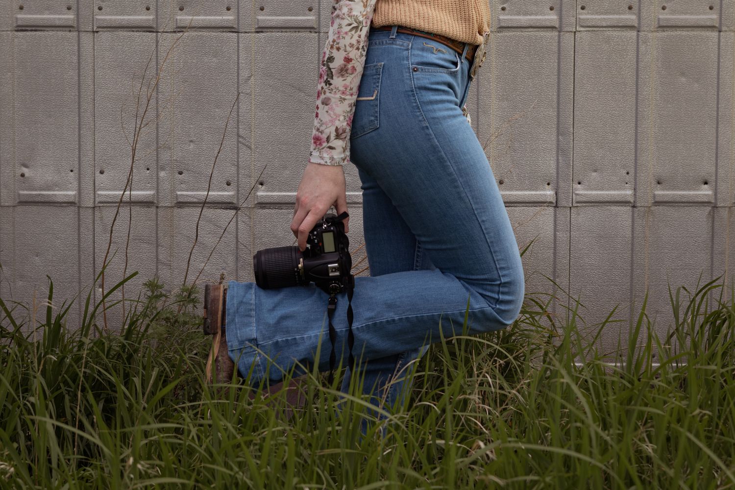 Woman in jeans holding a camera standing in tall grass against a concrete wall.