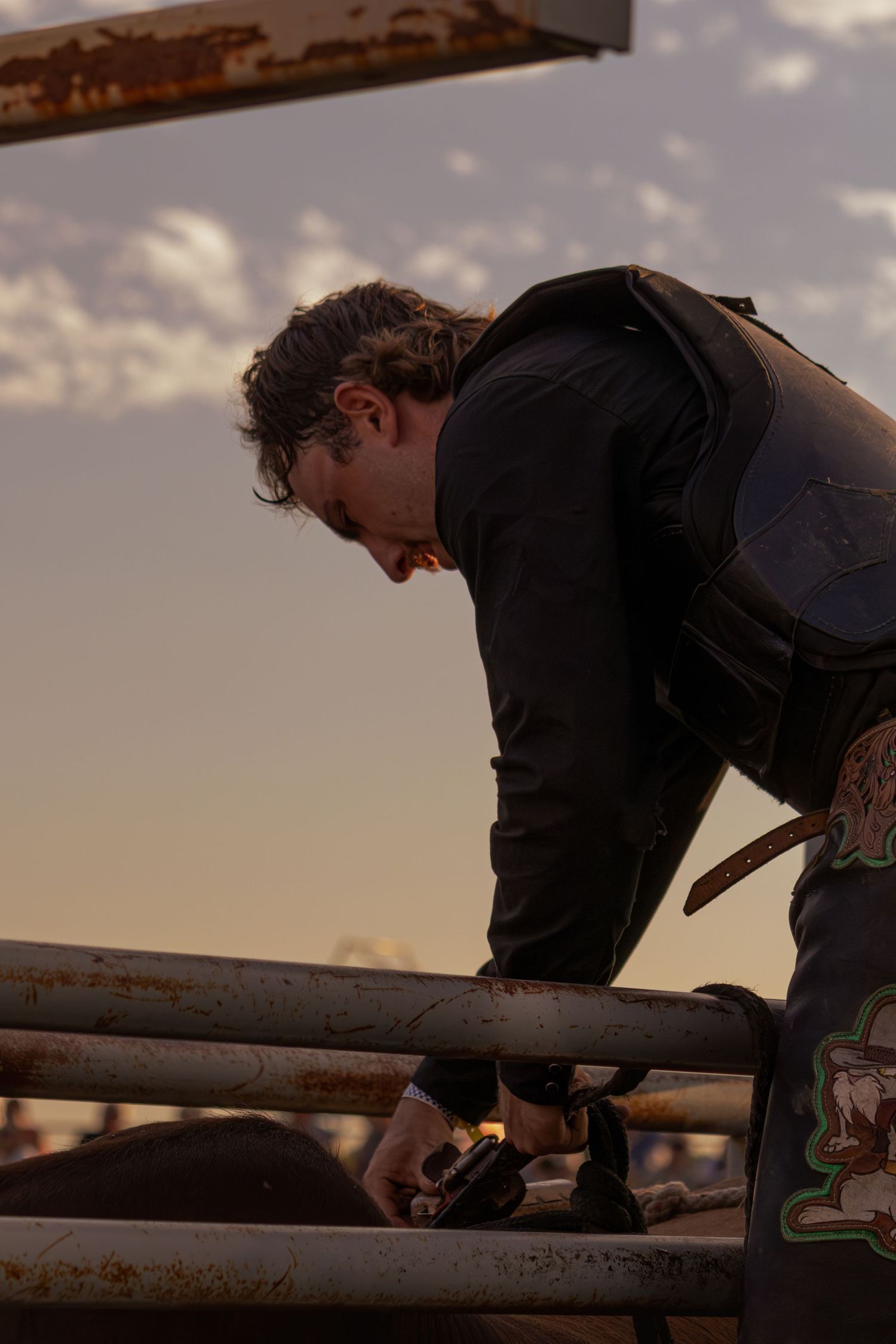 A cowboy in black leather vest leans over metal rodeo chute rails at sunset, preparing for a bull ride.