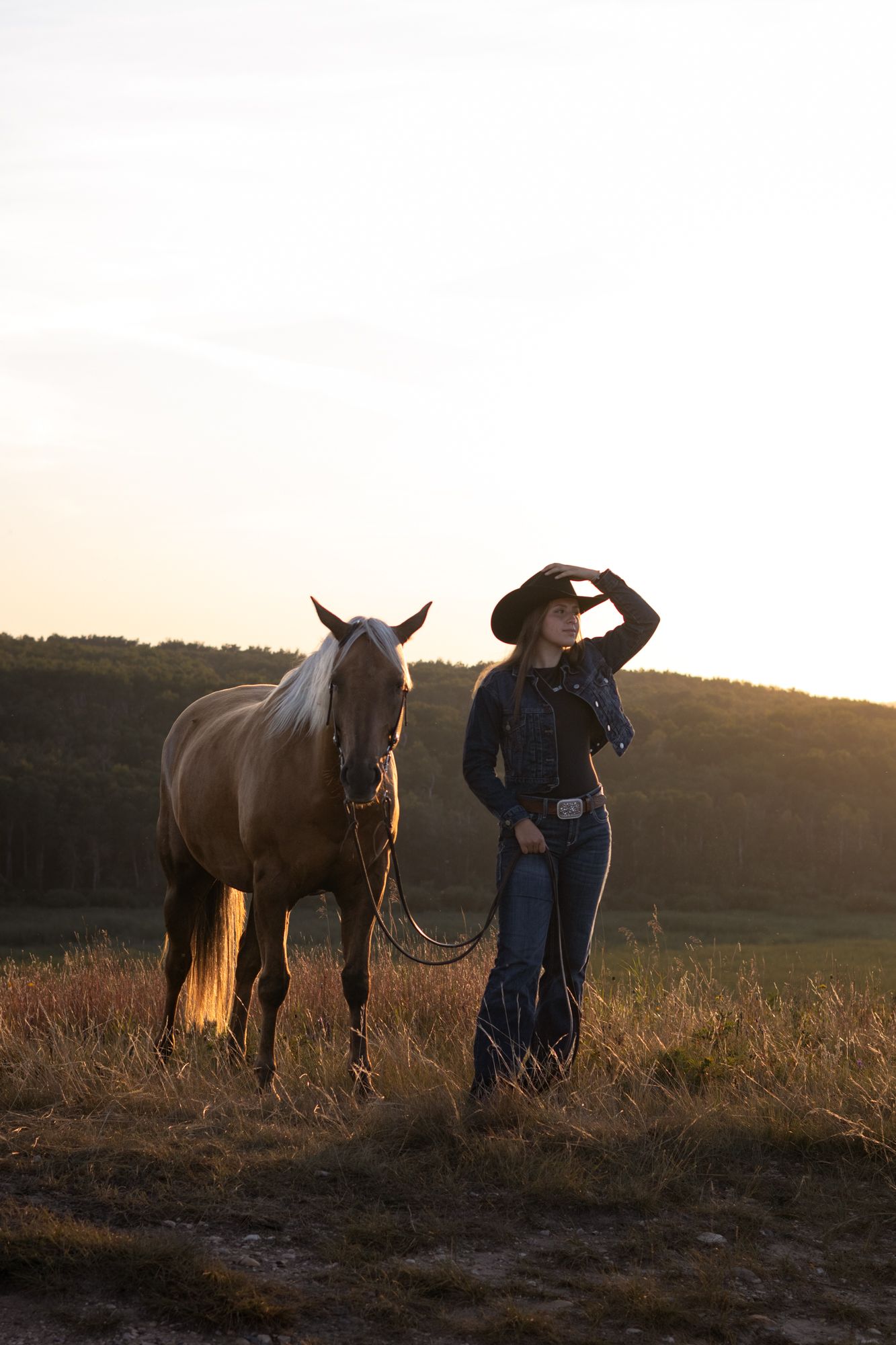 A cowgirl in a black hat and jeans stands with a palomino horse at golden sunset in an open field.
