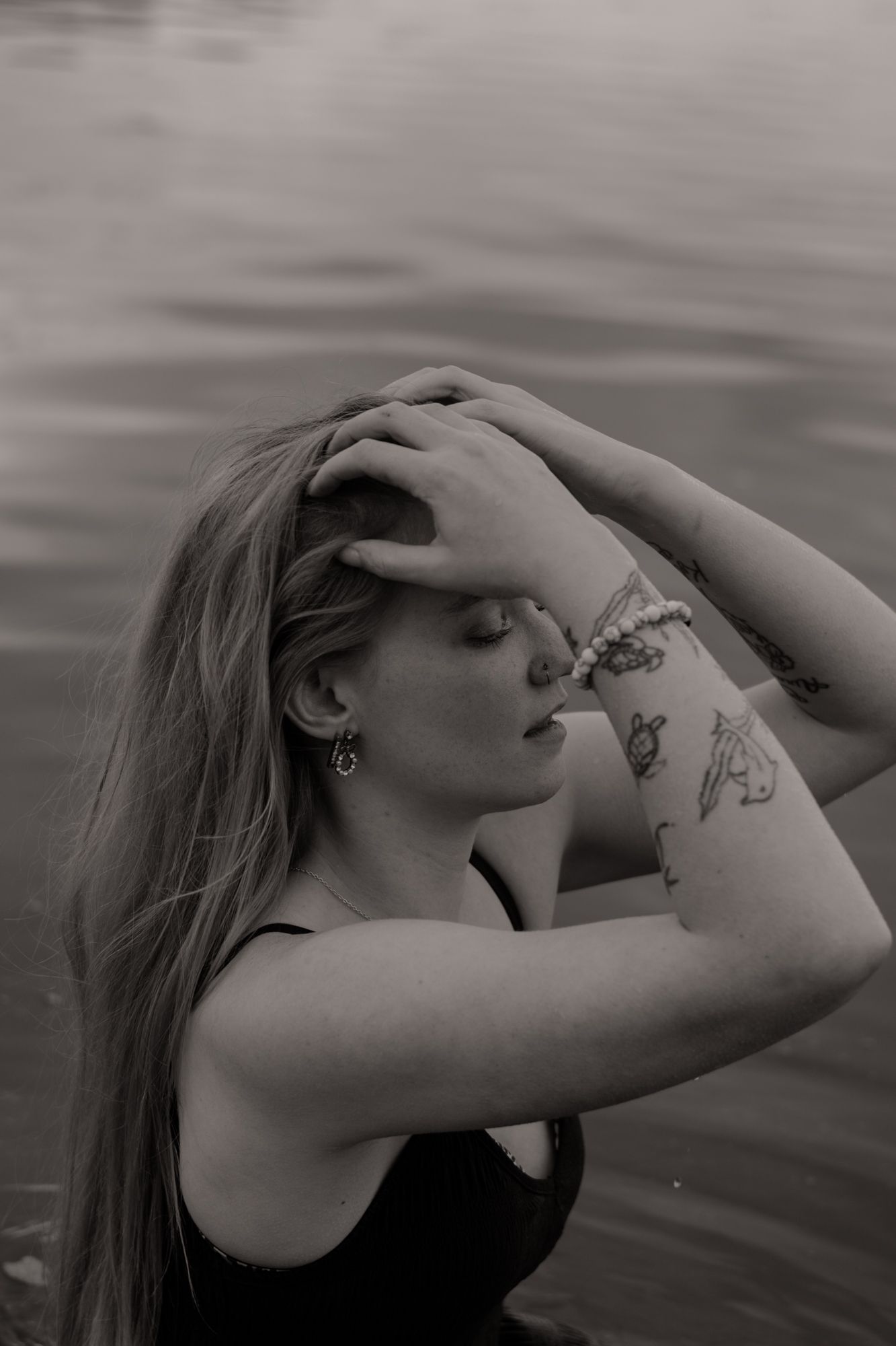 Black and white portrait of a young woman with tattoos and bracelet, hands in hair, eyes closed, near water.