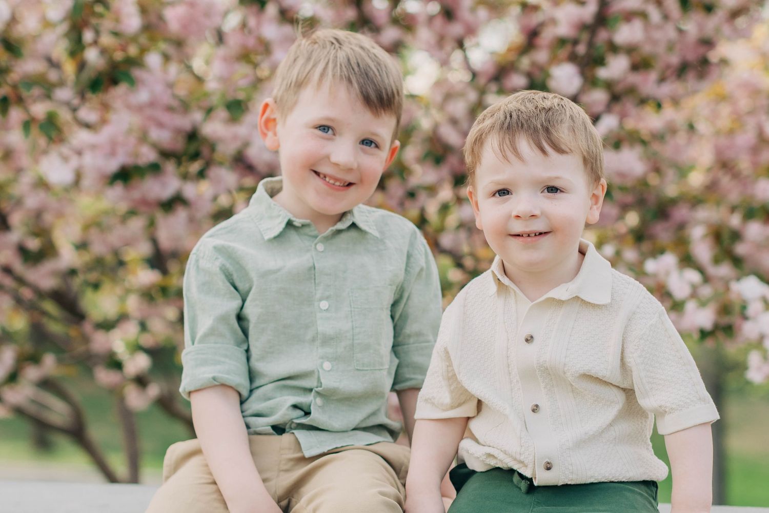 Two smiling young boys sitting together outdoors in front of pink cherry blossom trees on a sunny day.