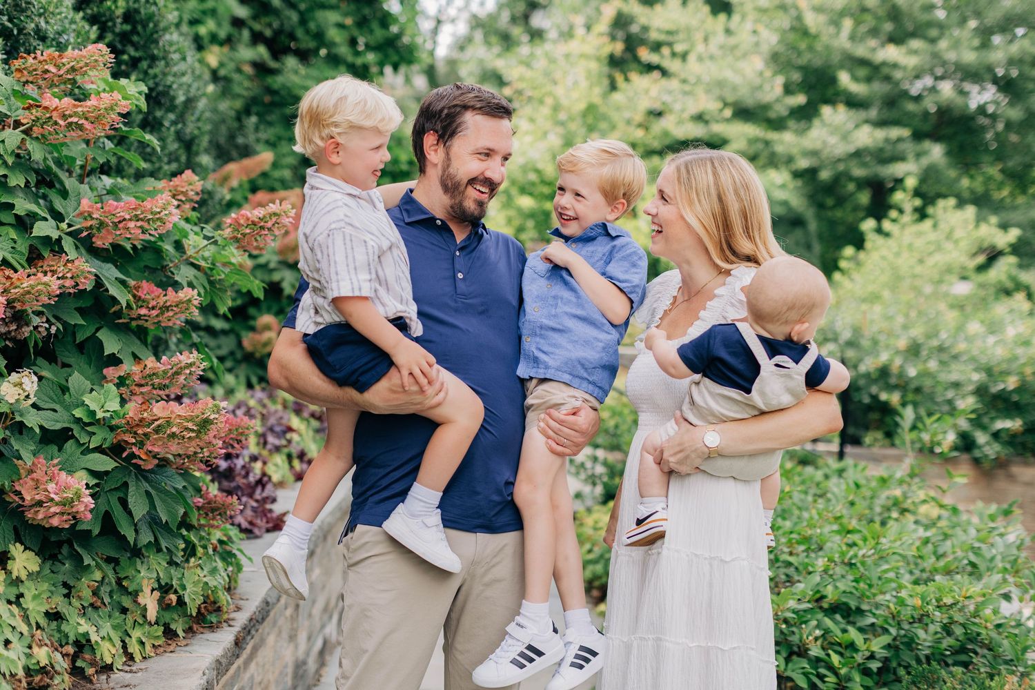 Happy family of four posing outdoors, parents holding two young boys, surrounded by lush green garden foliage.