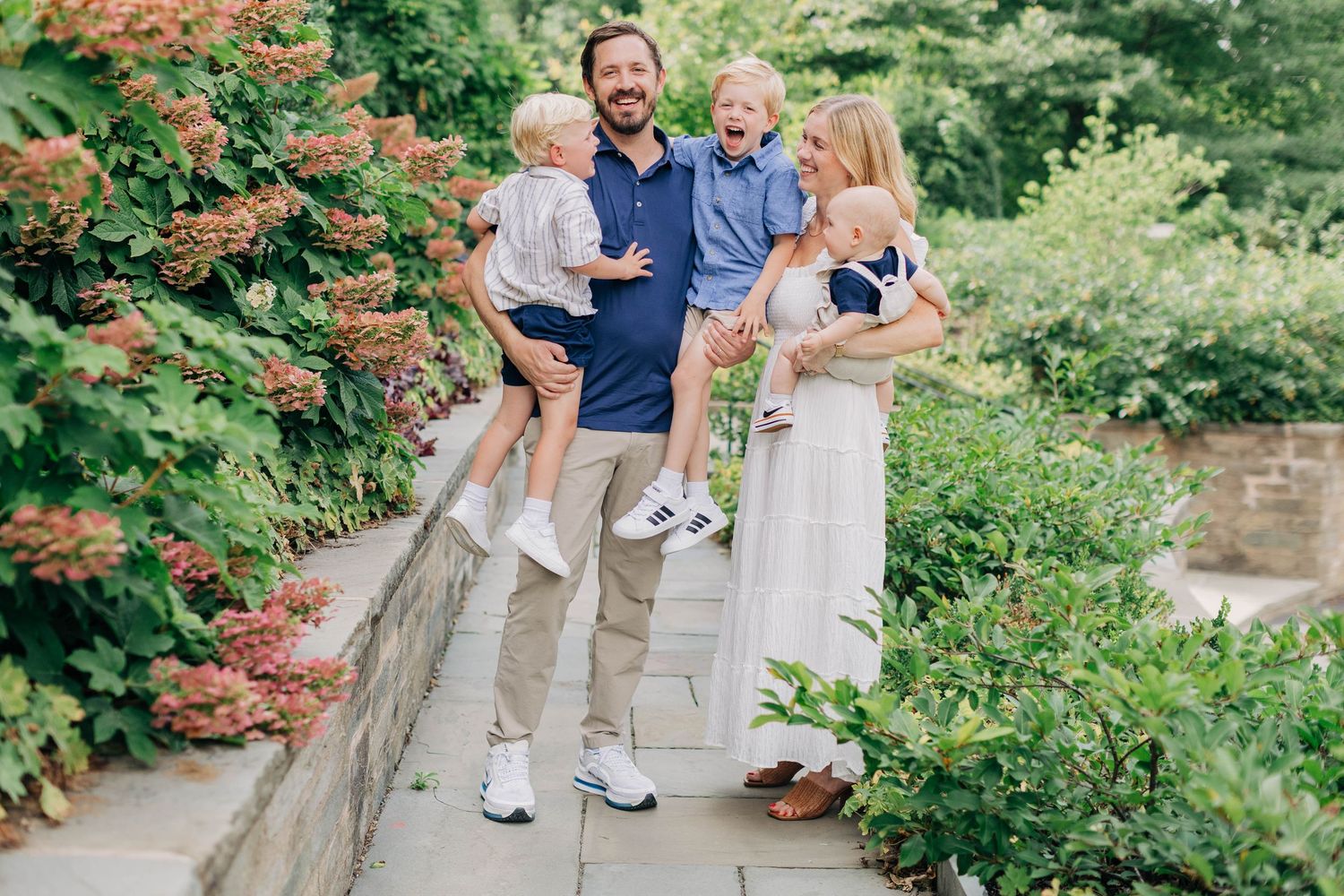 A happy family of four poses outdoors on a stone path surrounded by lush green gardens and blooming flowers.