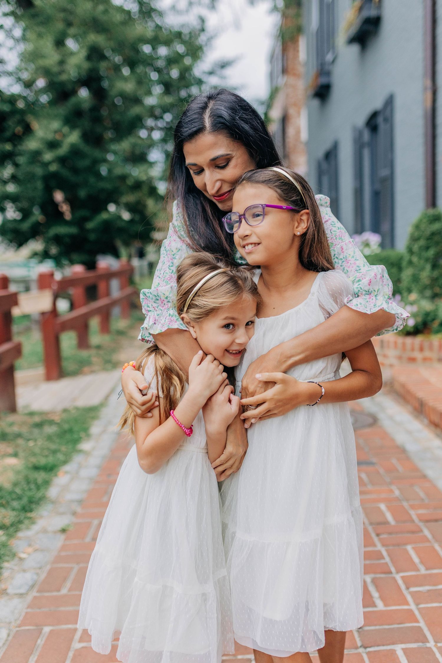 A mother tenderly hugs her two daughters in white dresses on a tree-lined path outdoors.