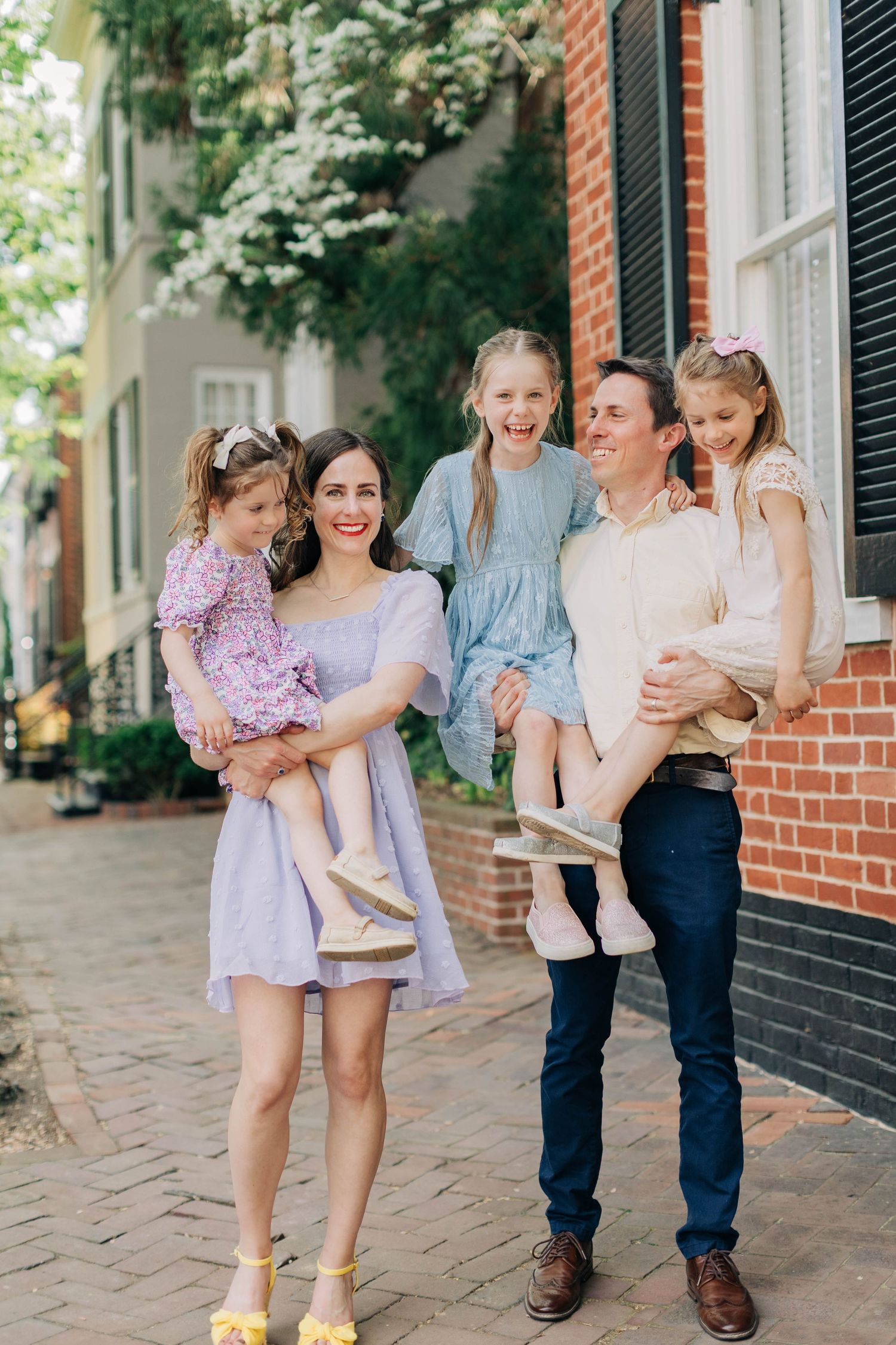 Smiling family of five on a brick pathway, parents holding three laughing girls in colorful spring outfits outdoors.