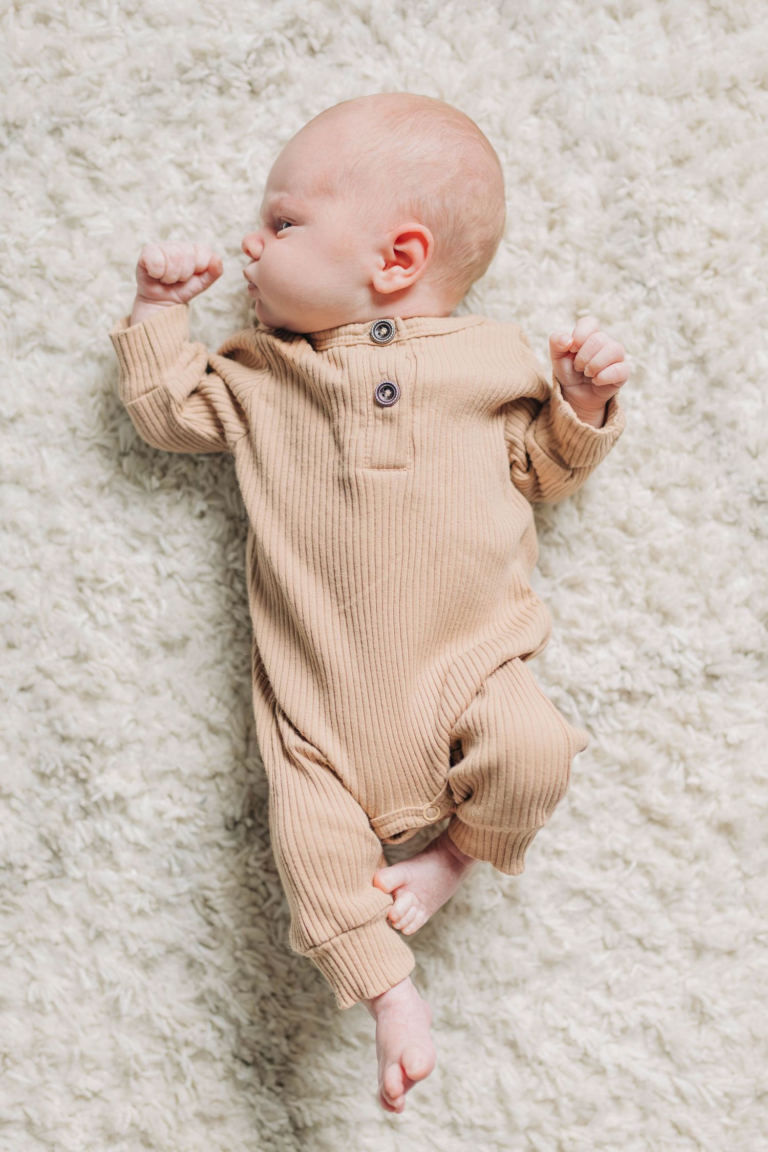 Baby wearing beige ribbed romper lying on white fluffy carpet.