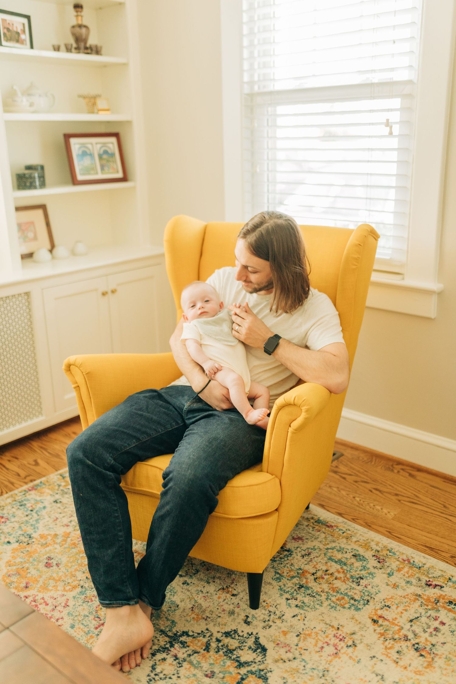 Family bonding moment in sunny living room with yellow armchair and floral rug.