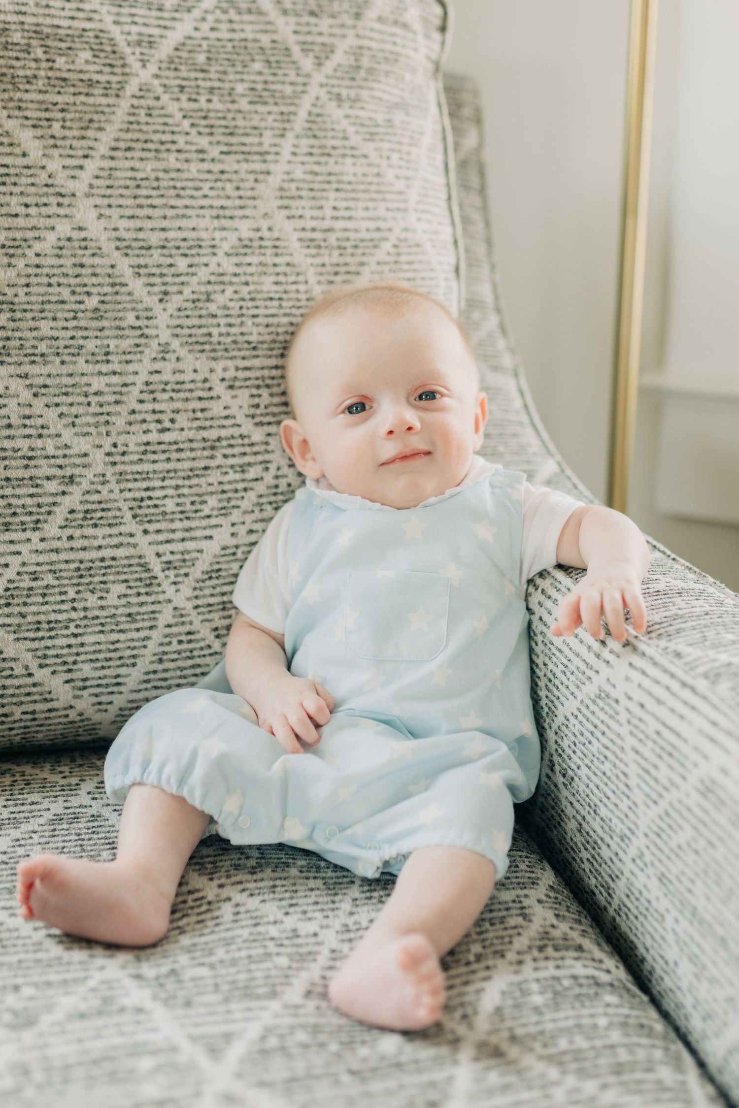 Infant in blue romper rests on gray patterned couch cushions.