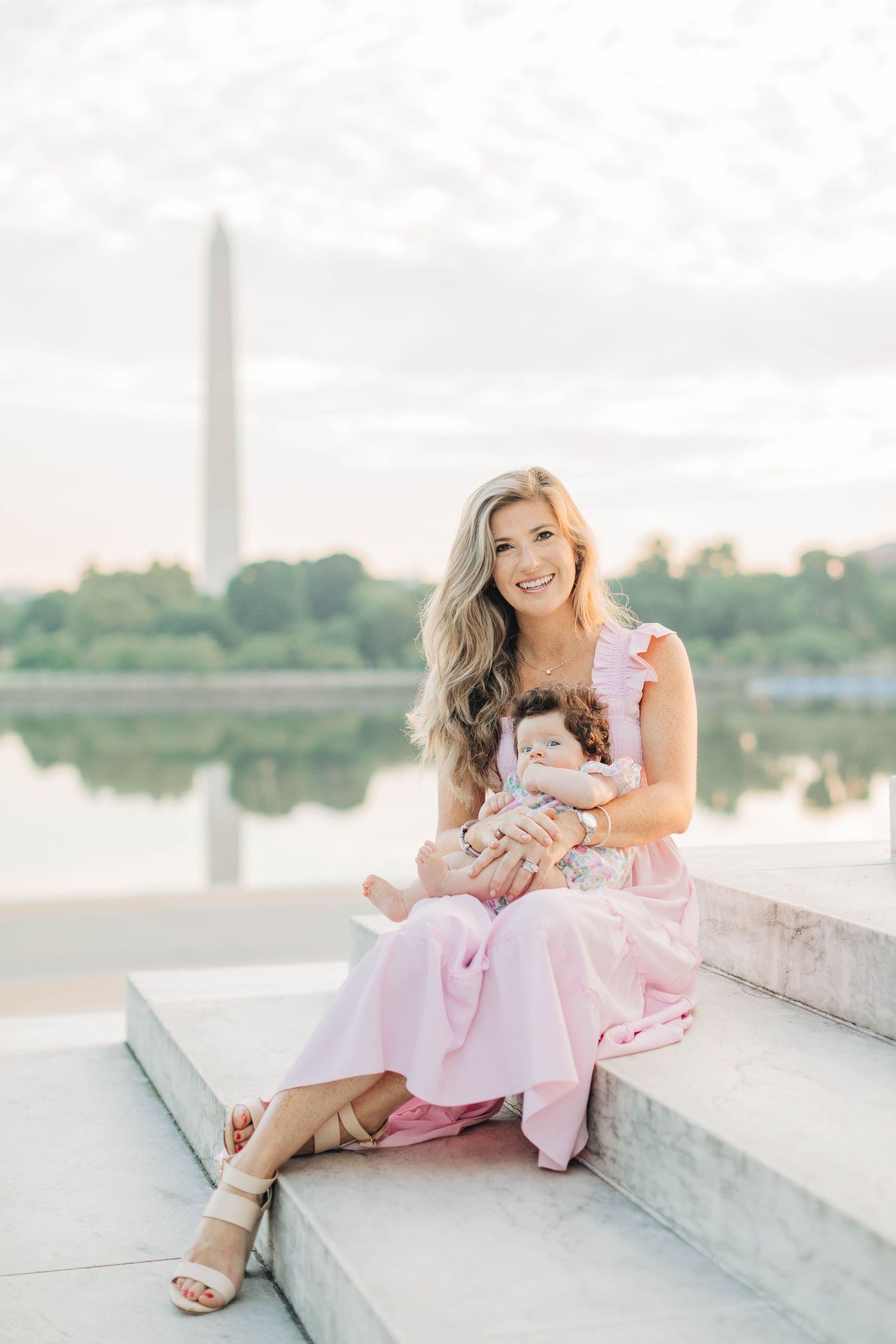 Woman in pink dress sitting on concrete steps near water at sunset.