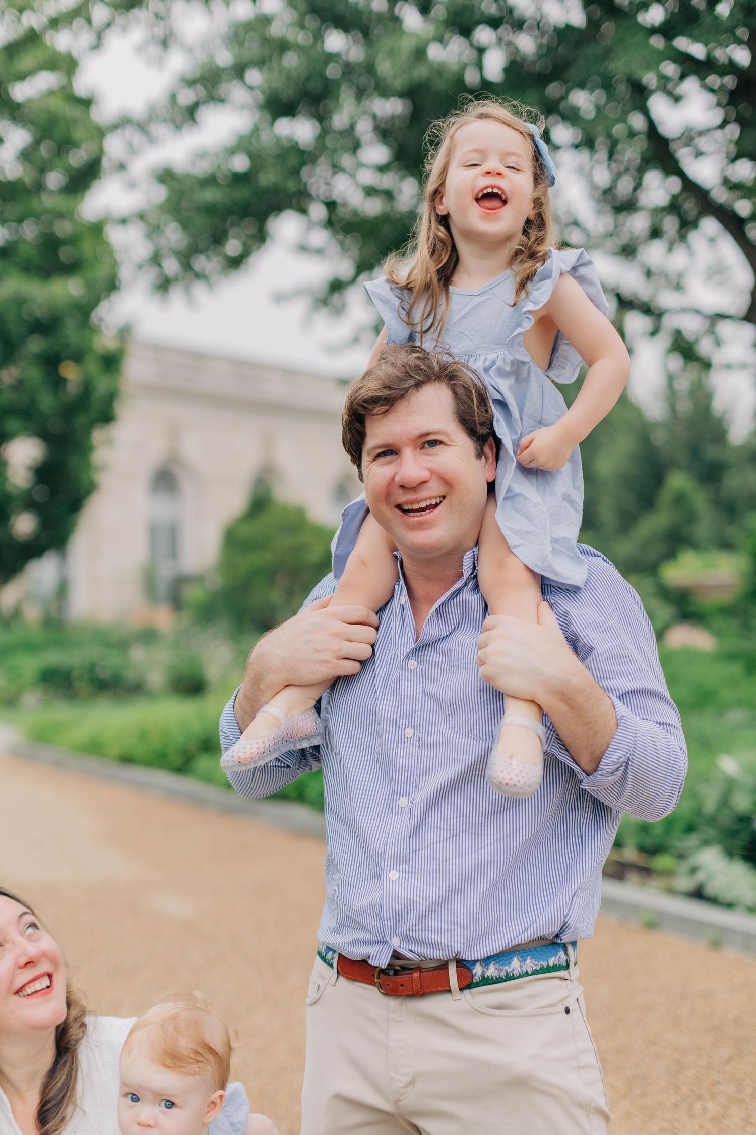 A laughing father carries his giggling toddler girl on his shoulders outdoors while a woman holds a baby nearby.