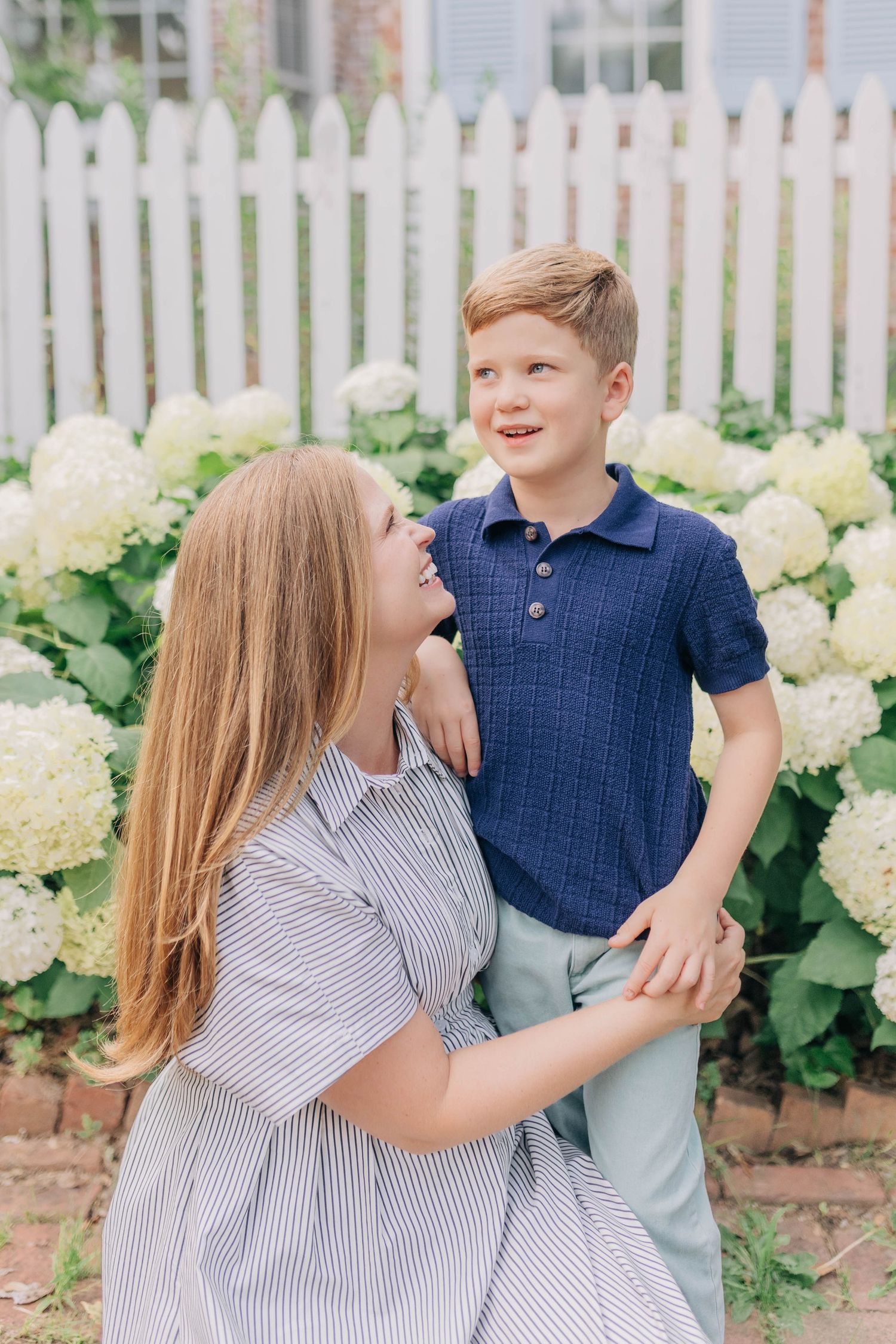 Mother crouching to embrace smiling red-haired boy near white fence with hydrangeas.
