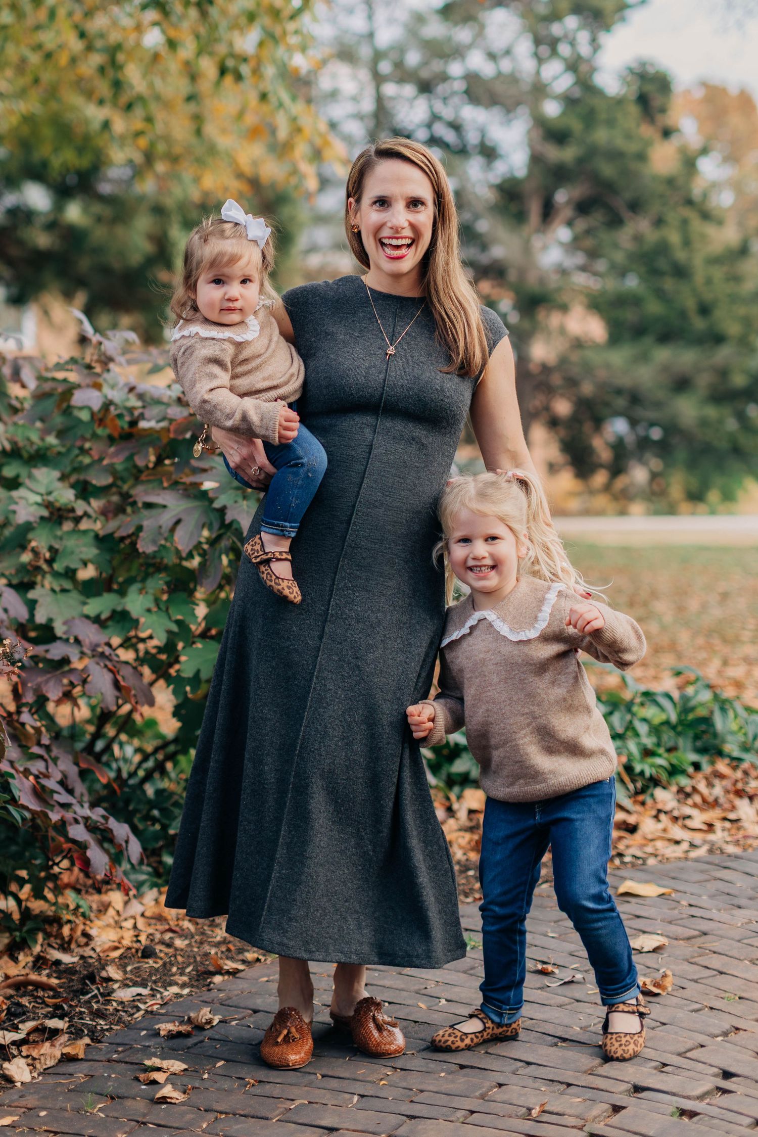 A smiling woman in a gray dress holds a toddler while a young girl in jeans stands beside her outdoors in autumn.