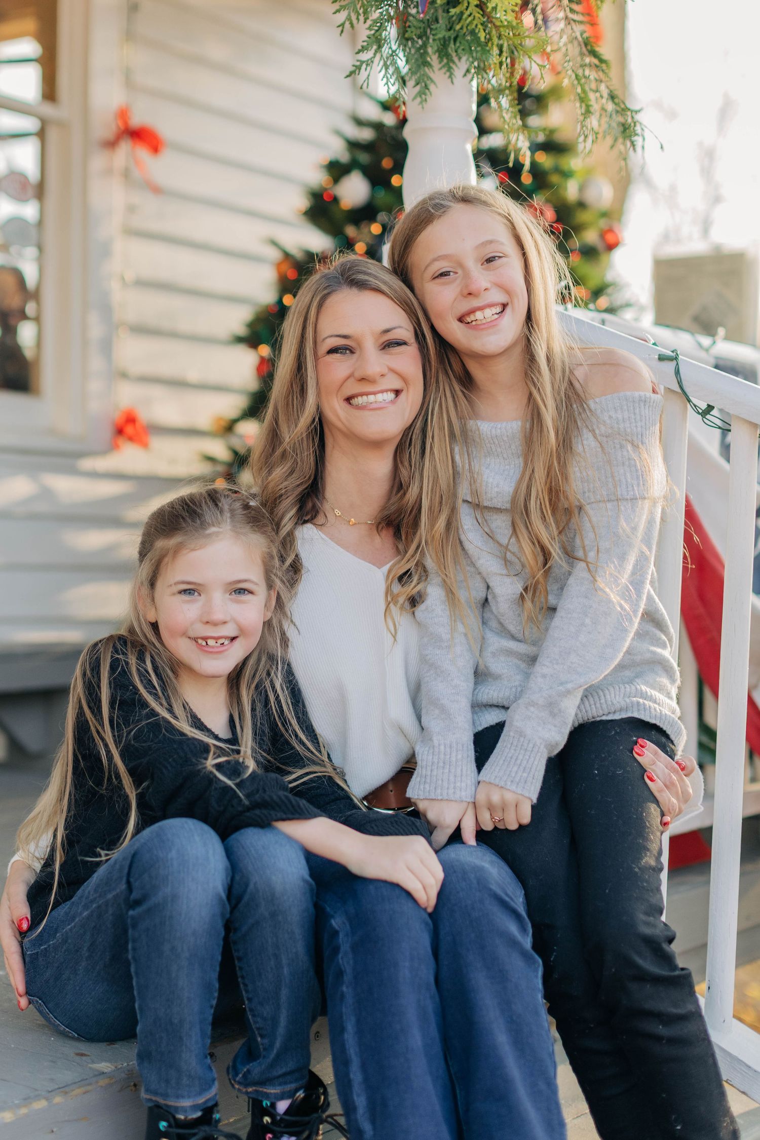 A woman and two girls smile together on porch steps decorated with a Christmas tree in the background.