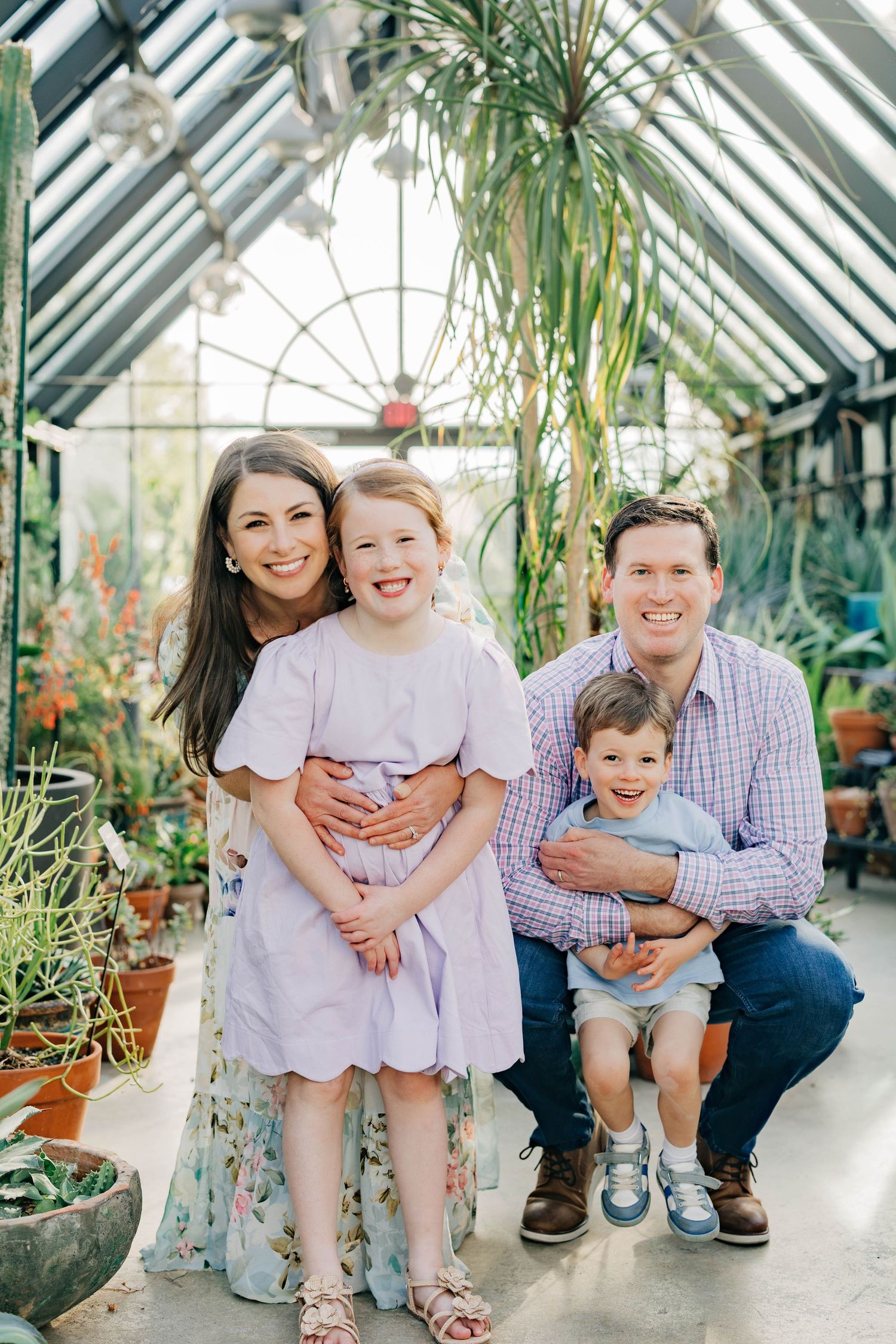 Family of four smiling in a lush greenhouse, parents with young daughter and son among tropical plants.