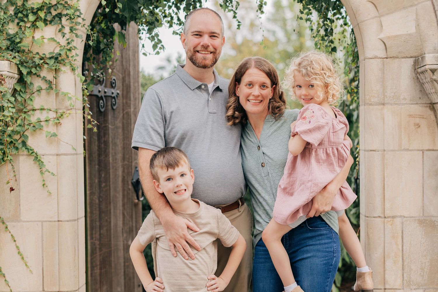 Smiling family of four with parents, a young boy, and a little girl posing together in front of a stone archway and wooden garden gate.