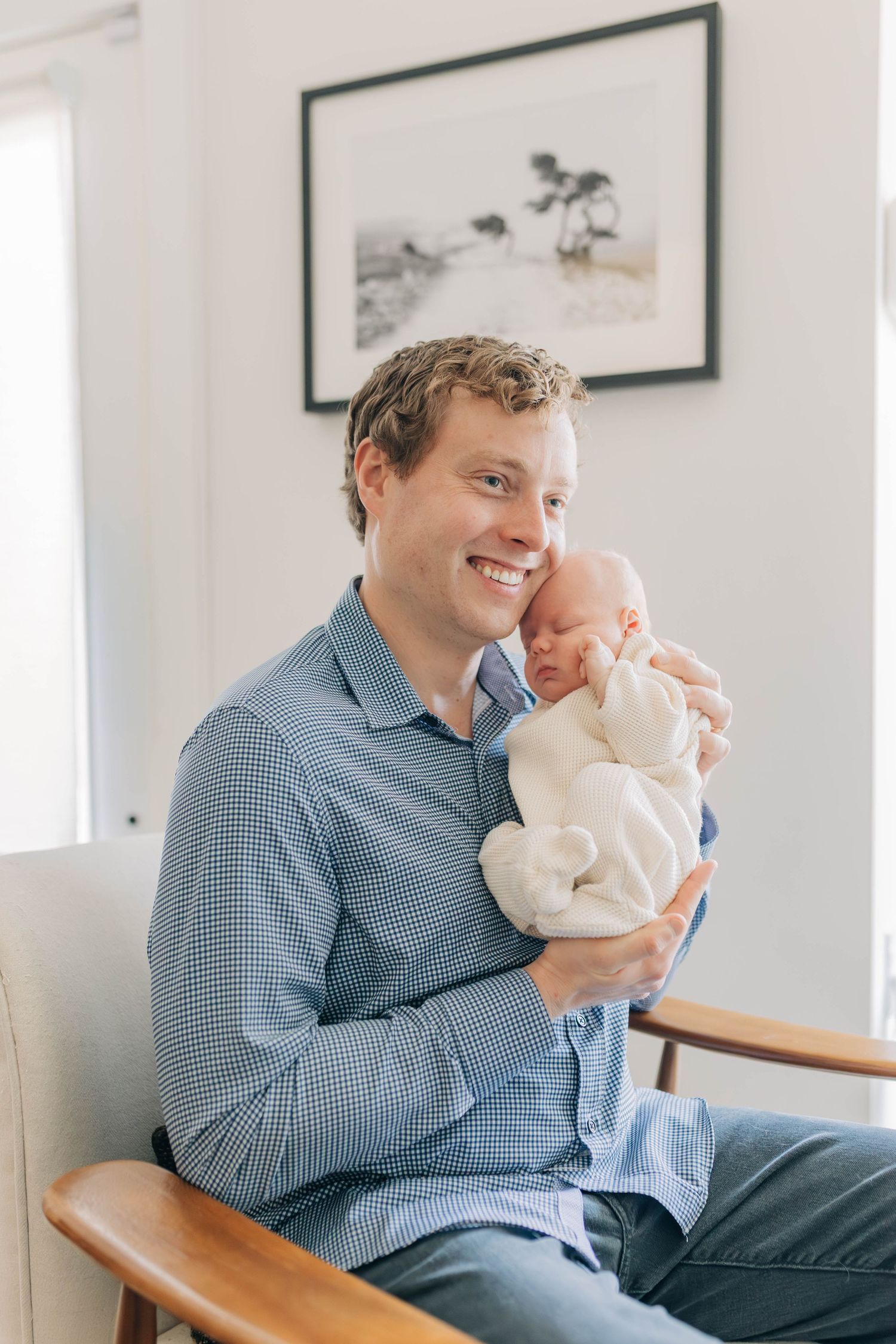 A smiling father holds his sleeping newborn baby close in a bright living room.