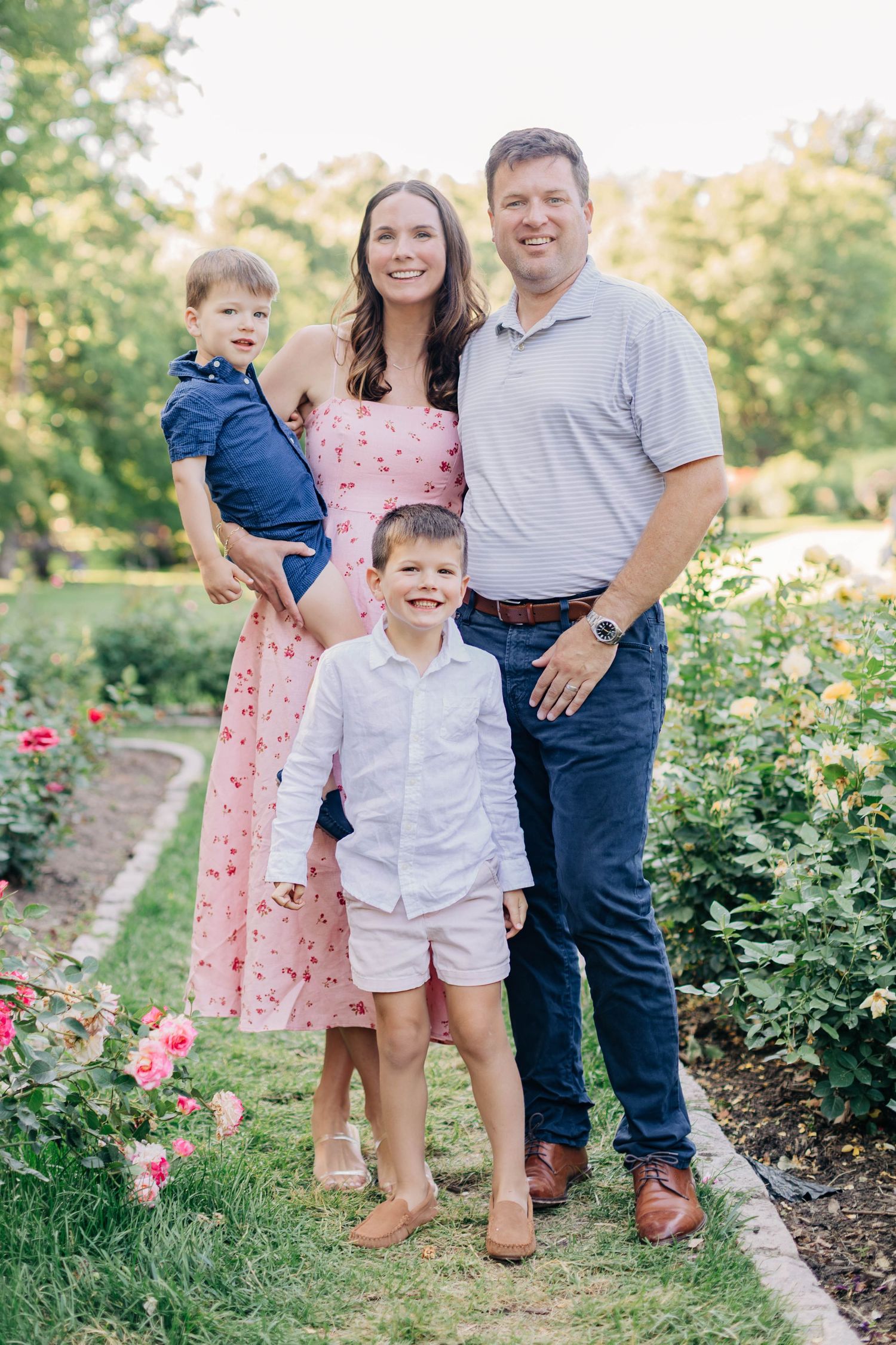 Family of four posing in a rose garden, mother in pink floral dress holding toddler, father and older son smiling outdoors.