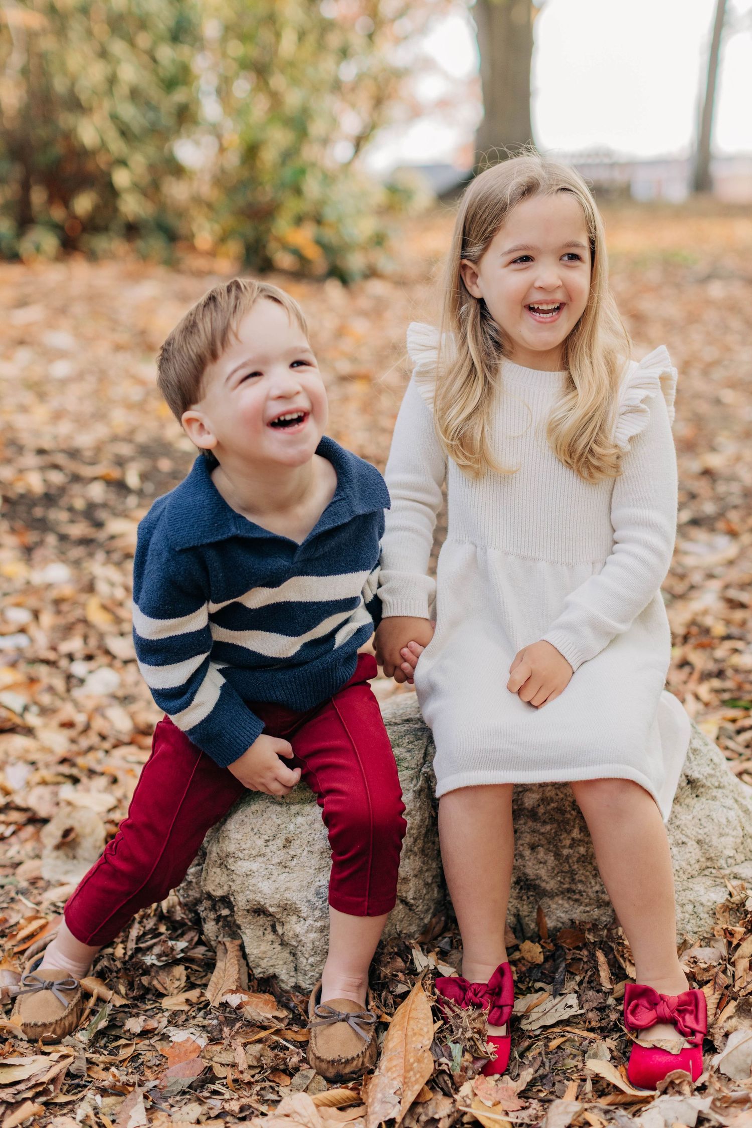 Two happy children sitting on a rock surrounded by autumn leaves, boy in striped sweater, girl in white dress.