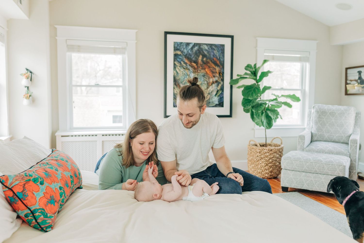 A family lounges together on a white bed in a bright room with houseplants and colorful pillows.