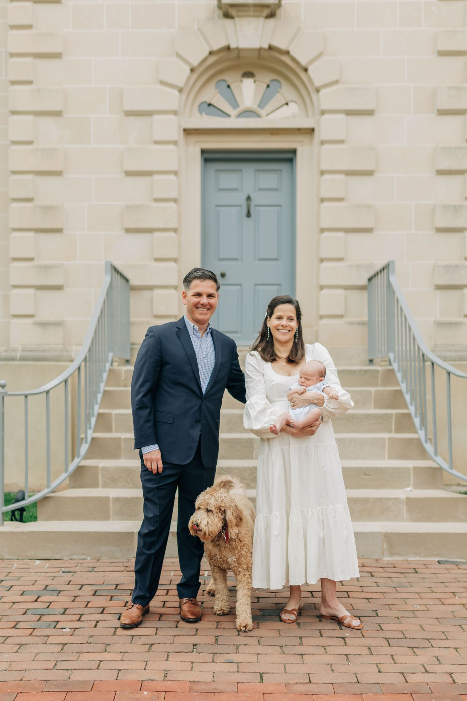 Family with baby and goldendoodle dog posing on brick steps of historic building with blue door.