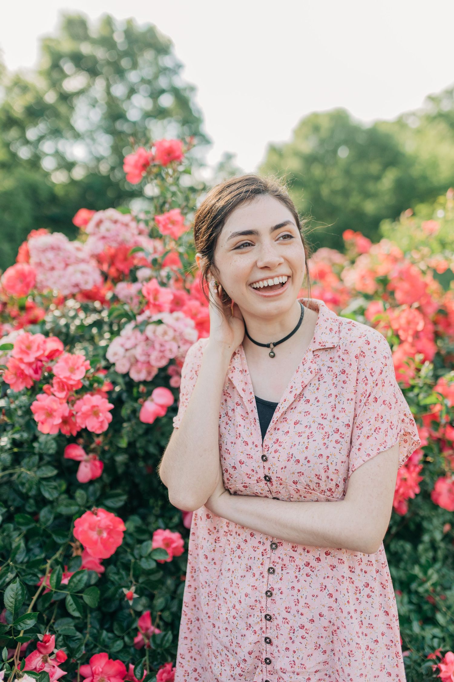 Person in pink floral dress poses playfully in front of blooming rose garden.