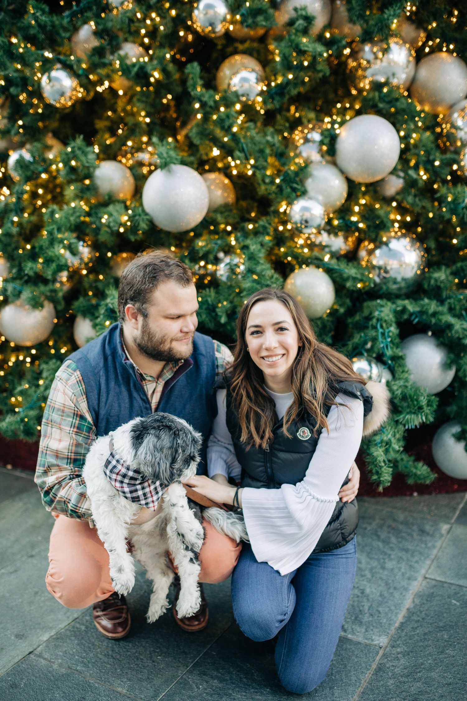 A couple smiles while posing with their dog in front of a festive Christmas tree.