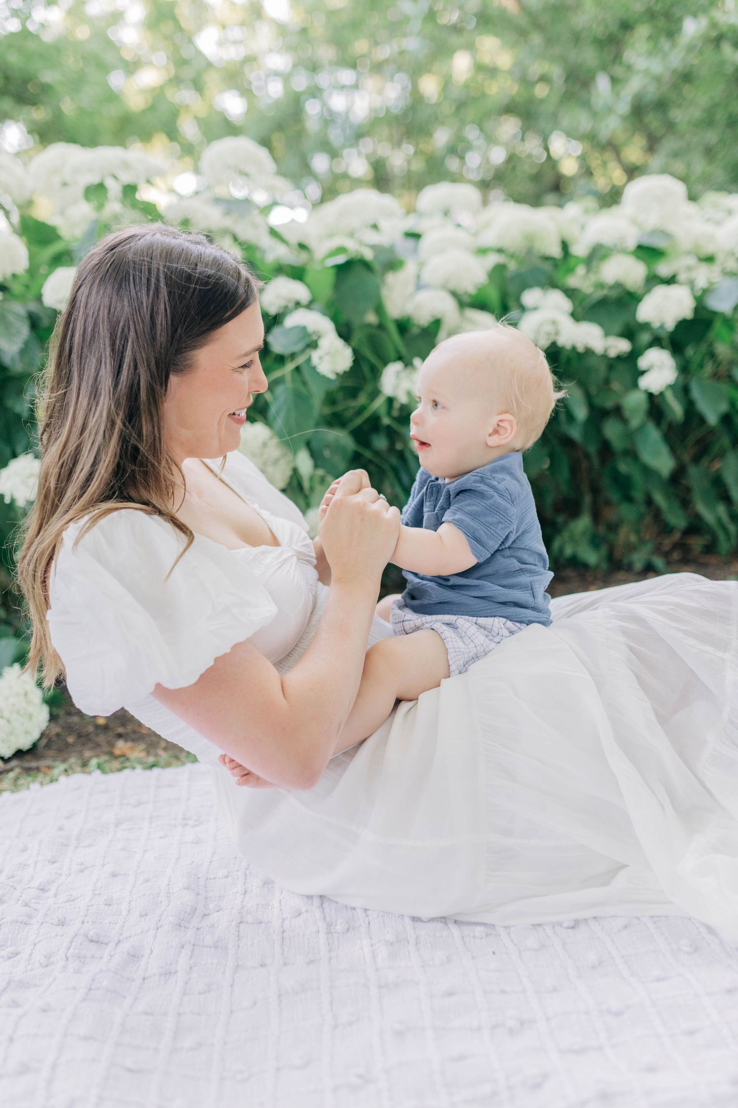 Tender moment between mother and baby on white blanket with flowering bushes in background.