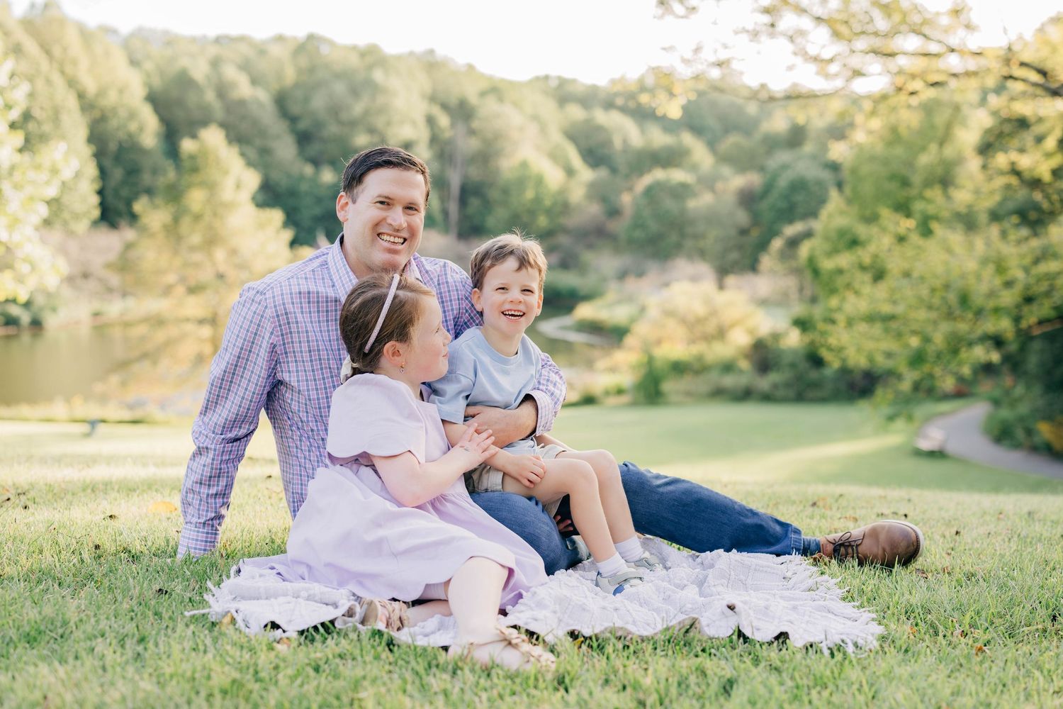 A smiling father sits on a blanket in a sunny park with his two young children, surrounded by lush green trees and a pond.