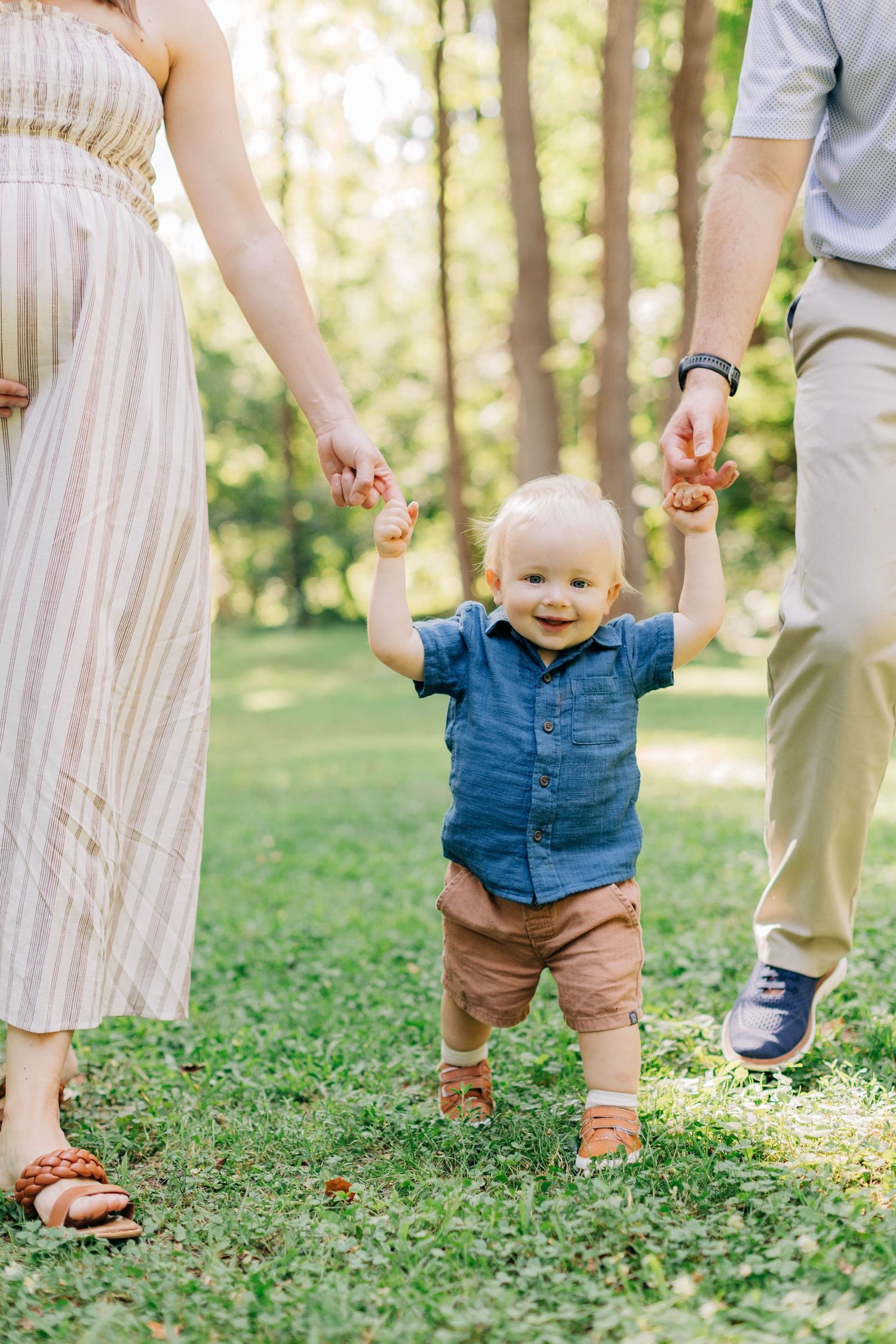 Smiling baby taking first steps outdoors, held by parents hands on green grass in sunny park.