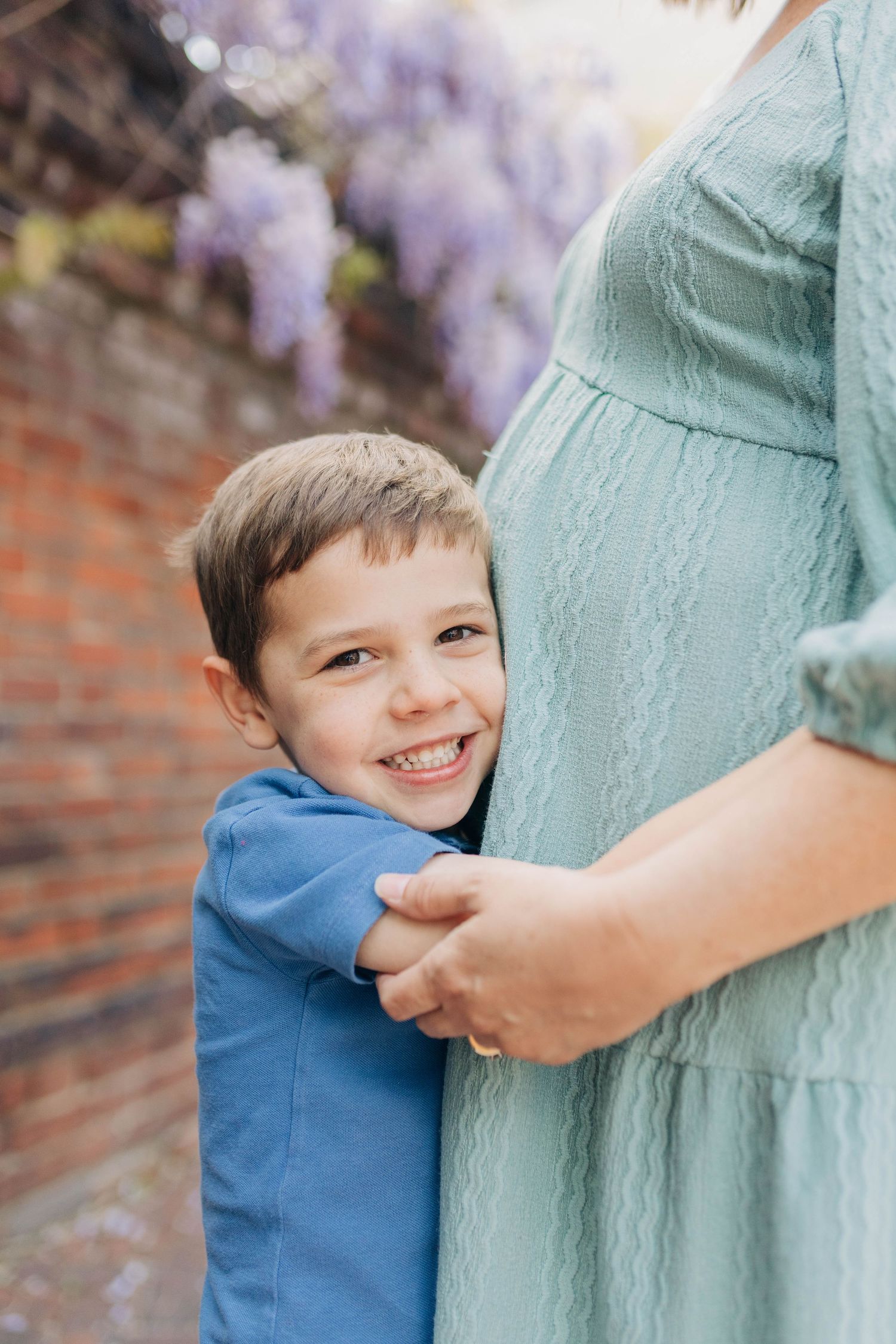 A smiling boy in a blue shirt hugs a pregnant woman in a mint dress outdoors near a brick wall with wisteria.