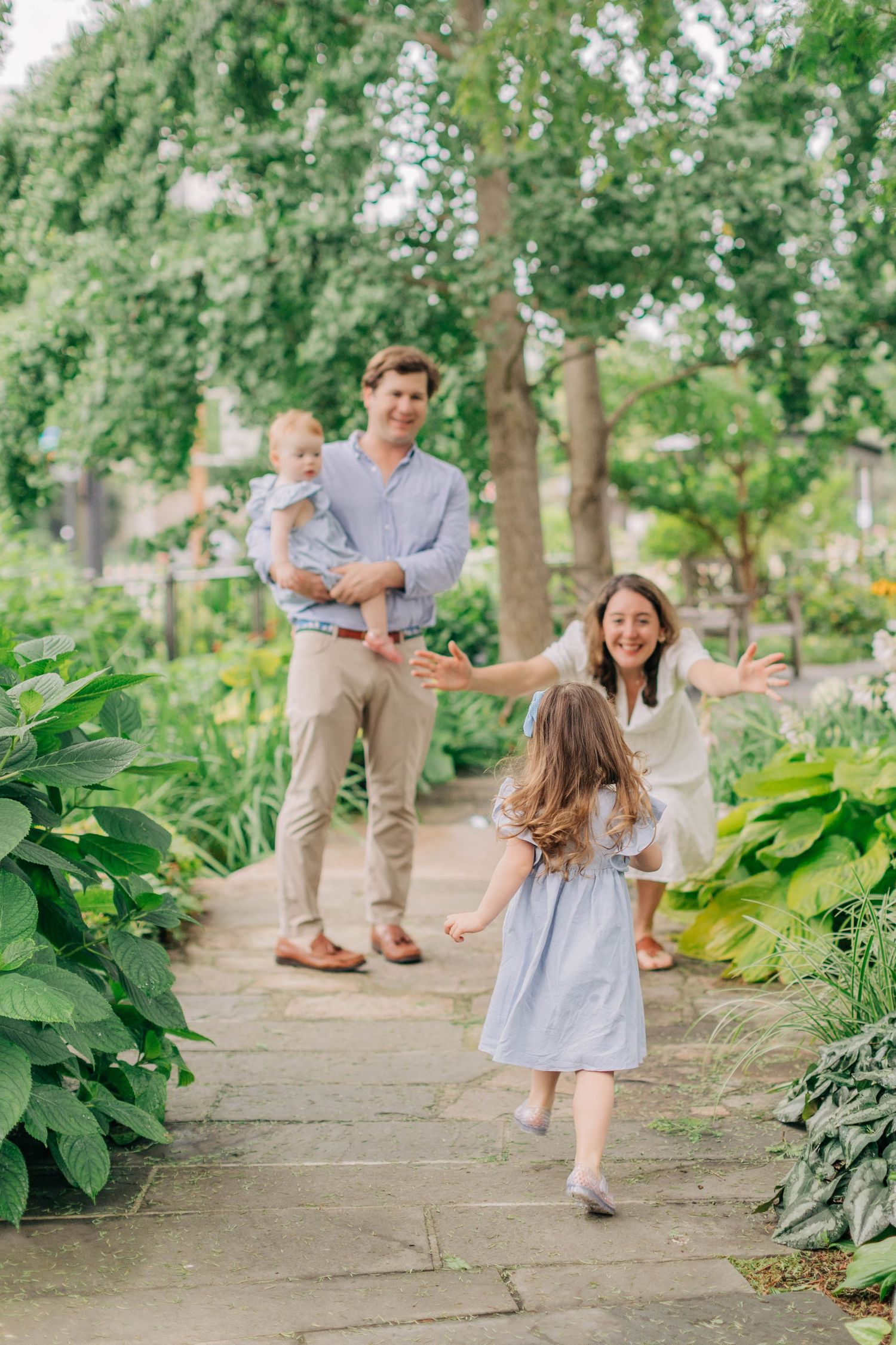 A joyful family reunion in a lush garden as a young girl runs toward her smiling parents and baby sibling.